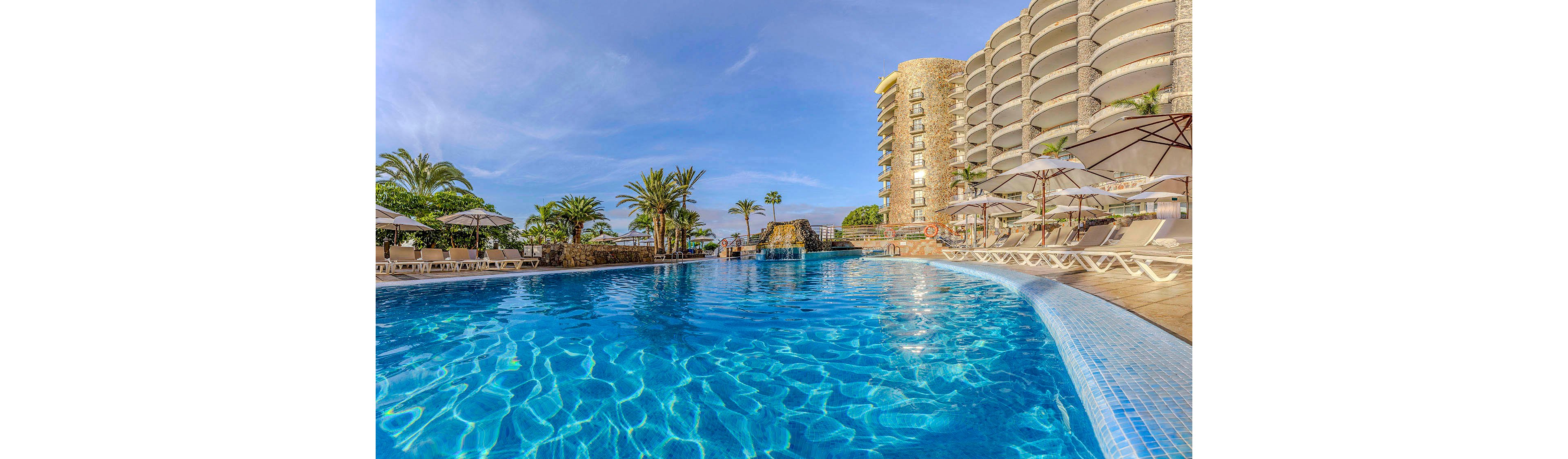A large, inviting outdoor swimming pool is surrounded by sun loungers and umbrellas at a modern hotel. The scene features clear blue water, palm trees, and a multi-story curved hotel building in the background. The setting is bright and sunny, evoking a relaxing vacation atmosphere. No visible text or numbers are present in the image.