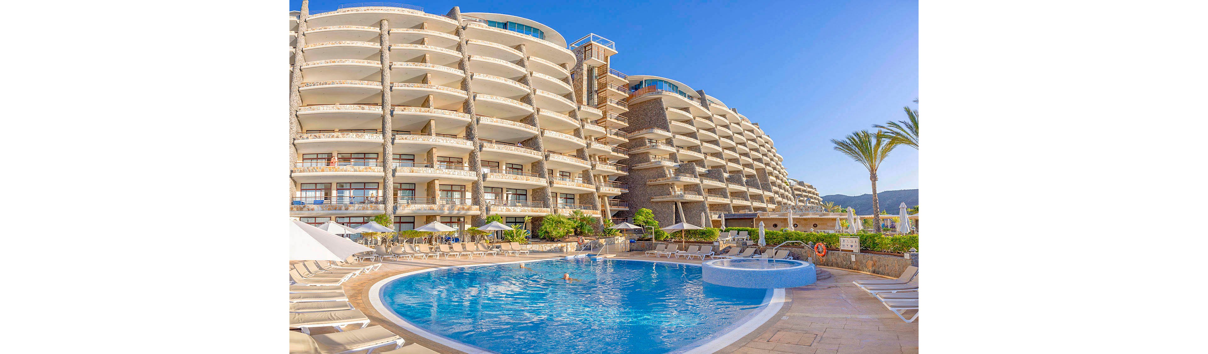 A large, multi-story resort hotel features curved balconies overlooking a spacious outdoor swimming pool. The pool area is surrounded by sun loungers, umbrellas, and palm trees, creating a relaxing vacation atmosphere. The sky is clear and blue, and a few people are enjoying the pool. The architecture is contemporary, with light-colored facades and glass railings. No visible text or numbers are present in the image.