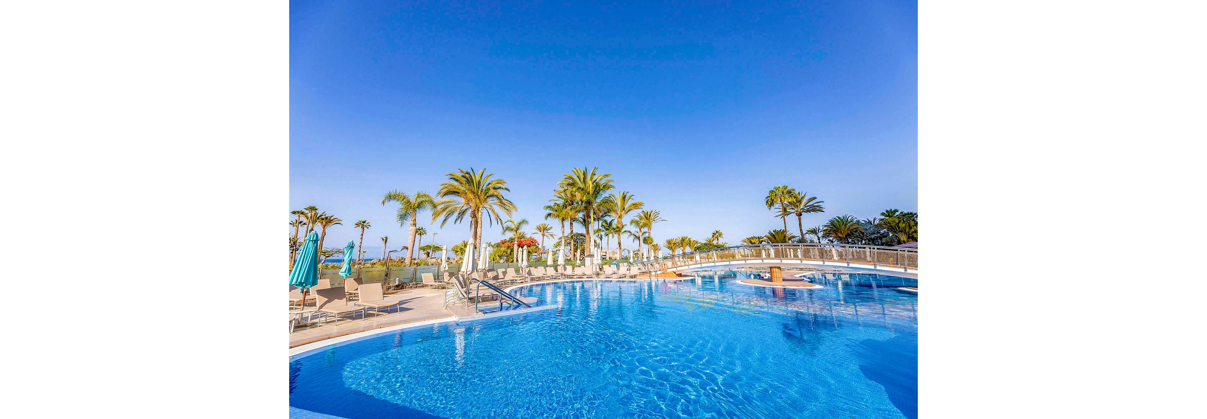 A large, inviting swimming pool is the main focus, surrounded by lounge chairs and umbrellas. Tall palm trees line the pool area, creating a tropical resort atmosphere. The sky is clear and blue, enhancing the bright and relaxing mood. No people are visible, emphasizing tranquility and leisure. The setting suggests a vacation or holiday destination.