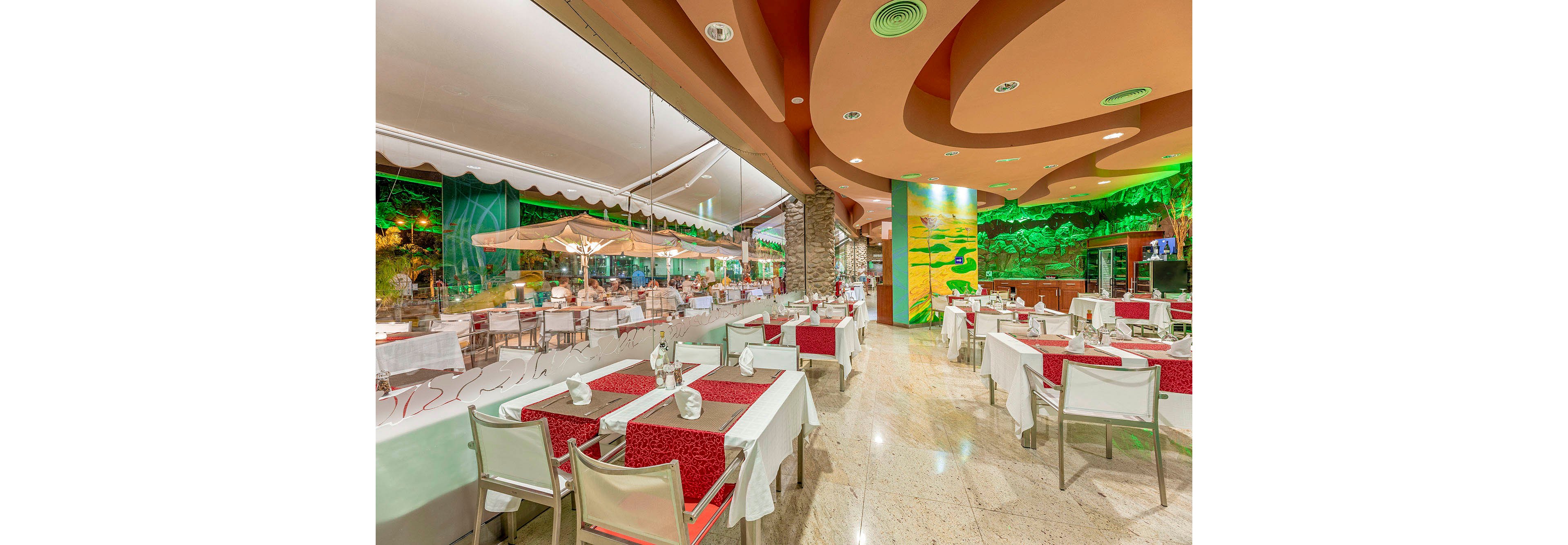 A spacious restaurant dining area features neatly arranged tables with red and white tablecloths and napkins. The setting includes a mix of indoor and outdoor seating, with large glass windows separating the spaces. The interior is brightly lit, with a colorful mural and contemporary ceiling design adding visual interest. Diners can be seen enjoying their meals in the background, creating a lively yet relaxed atmosphere.