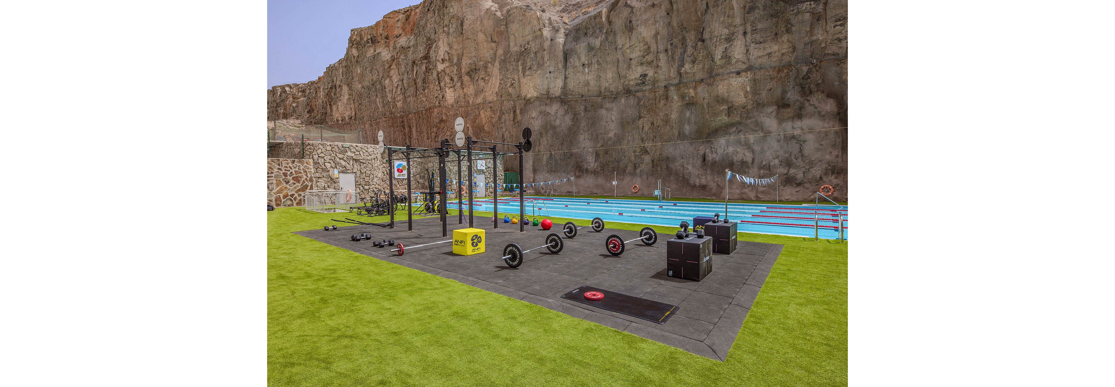An outdoor fitness area features various gym equipment including barbells, plyometric boxes, and medicine balls, set on rubber flooring. The gym is adjacent to a swimming pool with lane dividers, all situated against a dramatic rocky cliff backdrop. The setting is open-air with bright green artificial turf and clear skies, creating an energetic and active atmosphere. Visible numbers include '60' on a yellow plyometric box. No people are present in the scene.