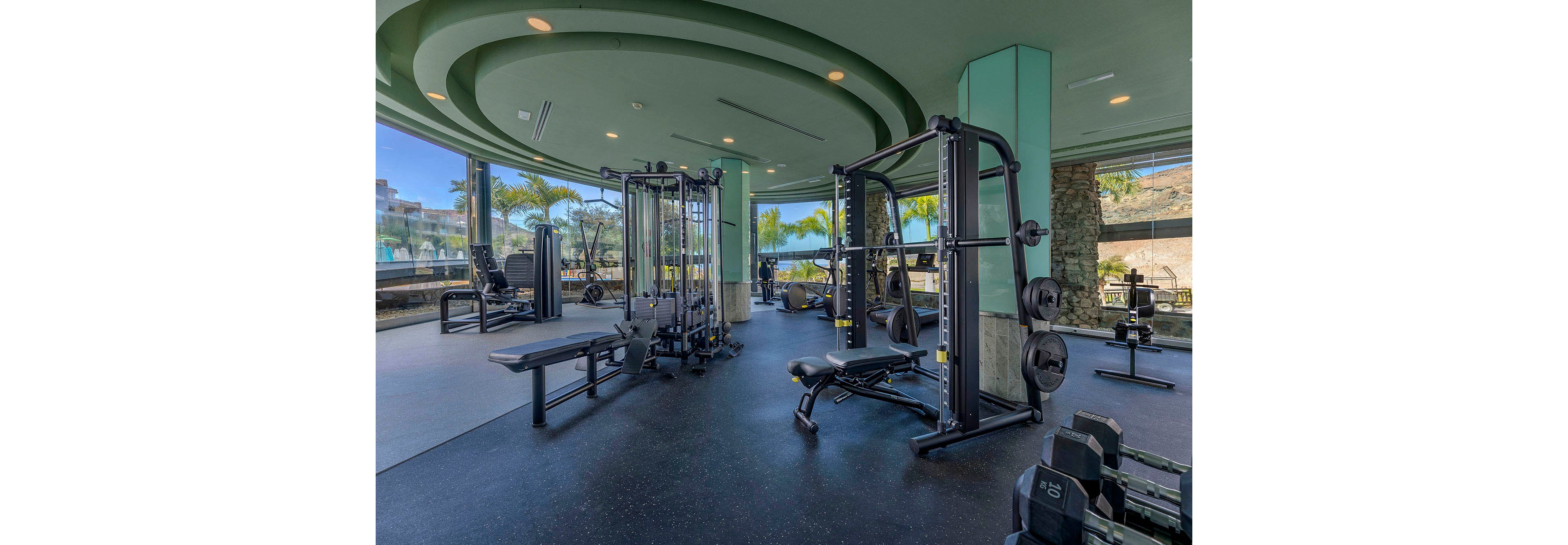 A spacious, contemporary gym features various weight machines and benches, set against large floor-to-ceiling windows. The setting is bright and airy, with palm trees and blue skies visible outside, suggesting a coastal or resort location. The interior design includes circular ceiling details and a clean, organized layout, creating an inviting atmosphere for fitness activities. No people are present, and no visible text or numbers appear on the equipment.