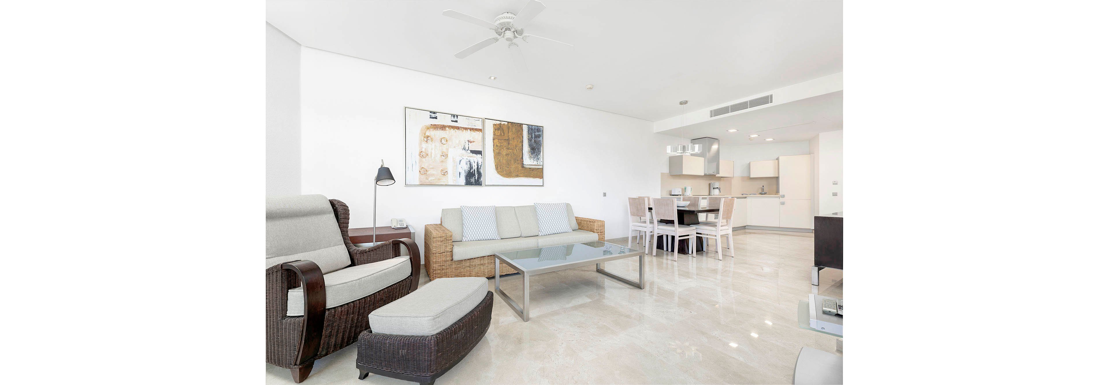 A spacious, contemporary living room features a wicker sofa with striped cushions, a matching armchair, and a glass coffee table. The open-plan layout connects to a dining area and a minimalist kitchen in the background. The room is decorated in neutral tones with abstract wall art, polished tile floors, and ample natural light. No visible text or numbers are present in the image.
