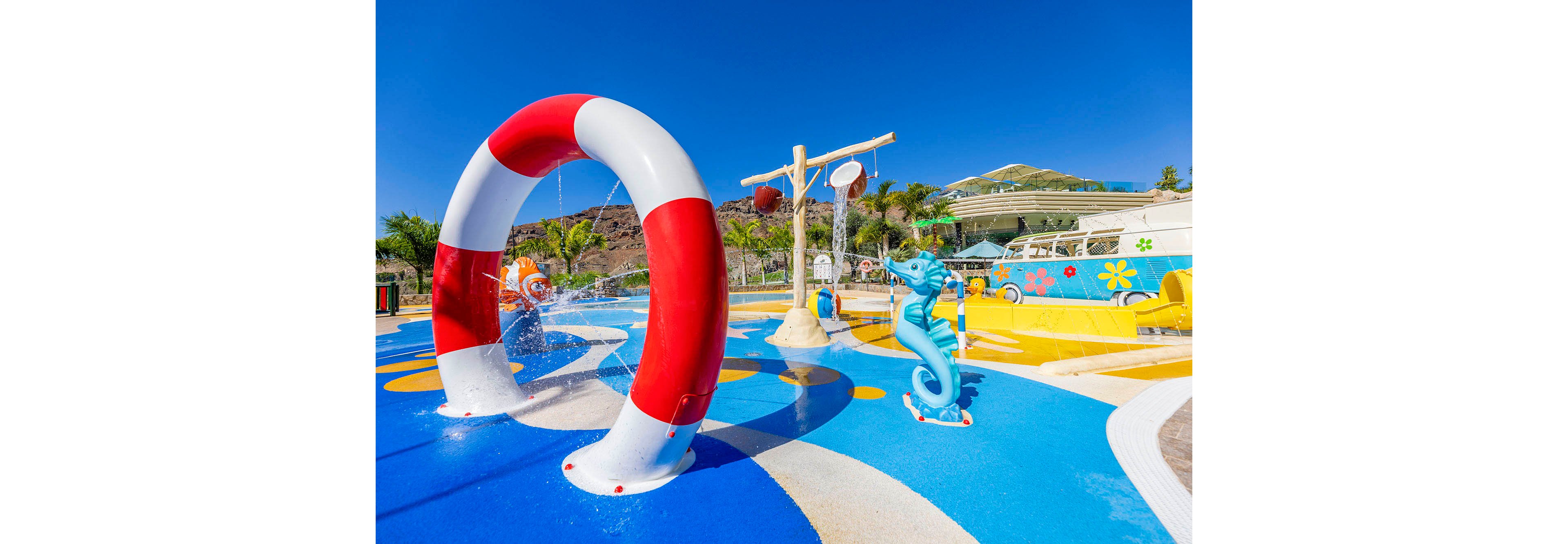 A vibrant splash pad features a large red and white arch spraying water, a blue seahorse sculpture, and interactive play elements. The setting is a sunny outdoor water park with palm trees and a bright blue sky. The ground is decorated with bold blue, yellow, and white patterns, creating a cheerful and inviting atmosphere. No visible text or numbers are present in the image.
