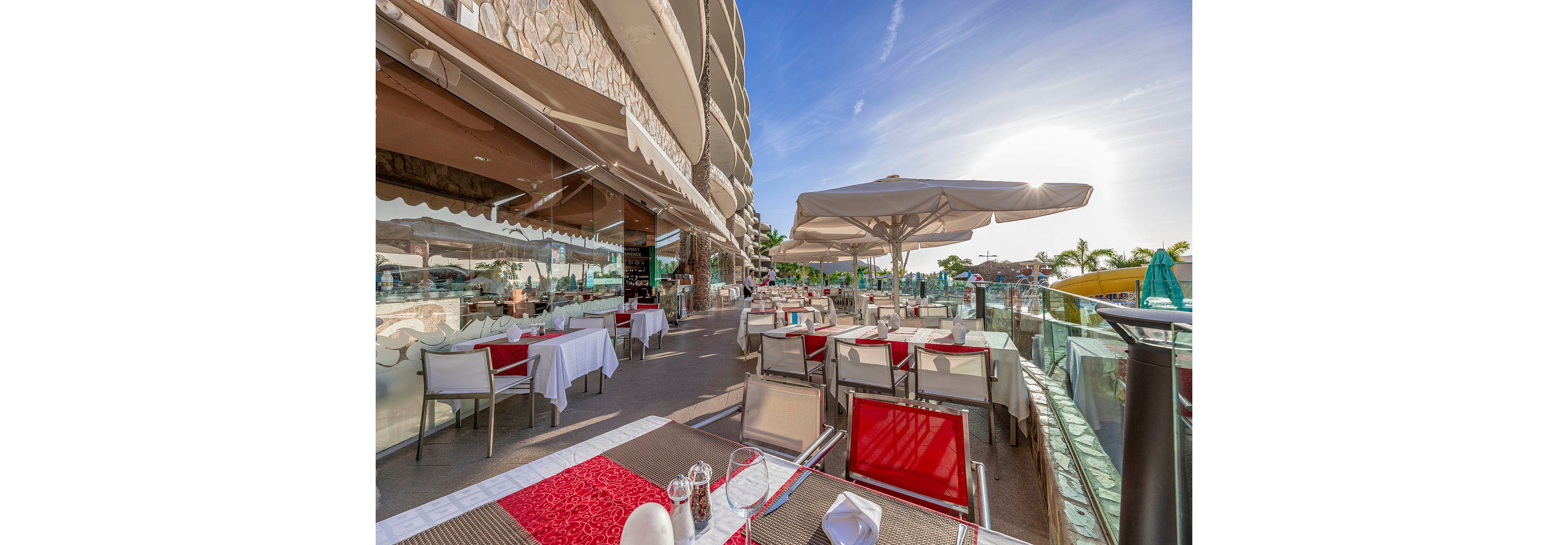 A spacious outdoor restaurant terrace features neatly arranged tables with red and beige accents, modern chairs, and large white umbrellas providing shade. The setting is adjacent to a contemporary building with glass railings and a view of palm trees in the distance. The scene is brightly lit by the afternoon sun, creating a welcoming and relaxed atmosphere for dining.