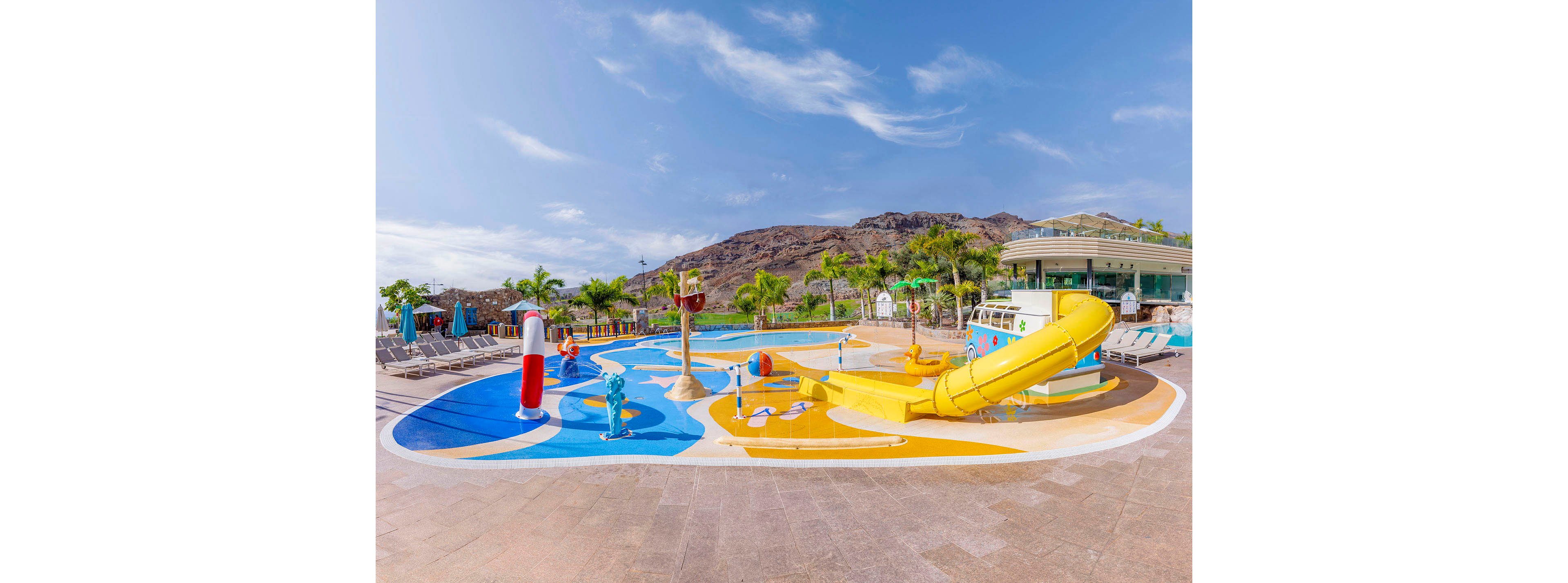 A vibrant children's water playground features a yellow slide, splash pads, and playful water structures. The setting is an outdoor pool area surrounded by palm trees and lounge chairs, with mountains visible in the background. The scene is bright and sunny, evoking a cheerful and energetic mood. No visible text or numbers are present in the image.