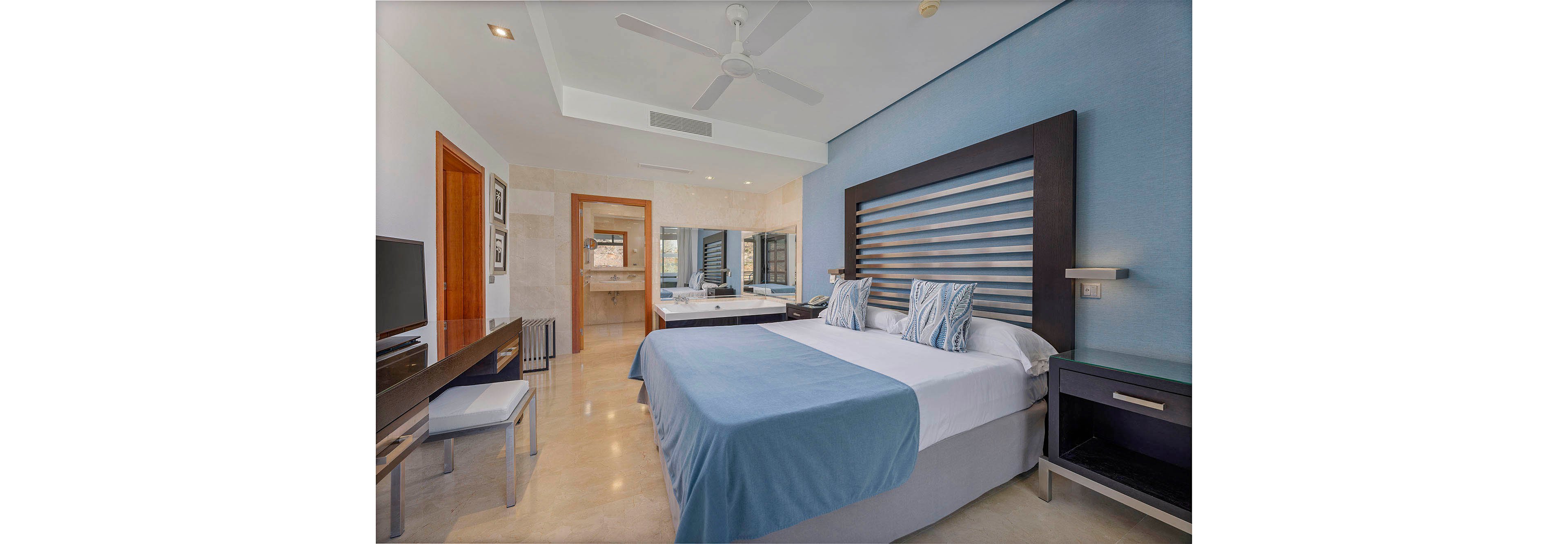 A contemporary hotel room features a large bed with blue and white linens, complemented by patterned pillows. The space includes a sleek desk, a wall-mounted mirror, and a visible bathroom area with a double sink. The room is well-lit with natural light, and the overall palette is cool and calming with blue and neutral tones. Minimalist furniture and clean lines enhance the modern aesthetic.