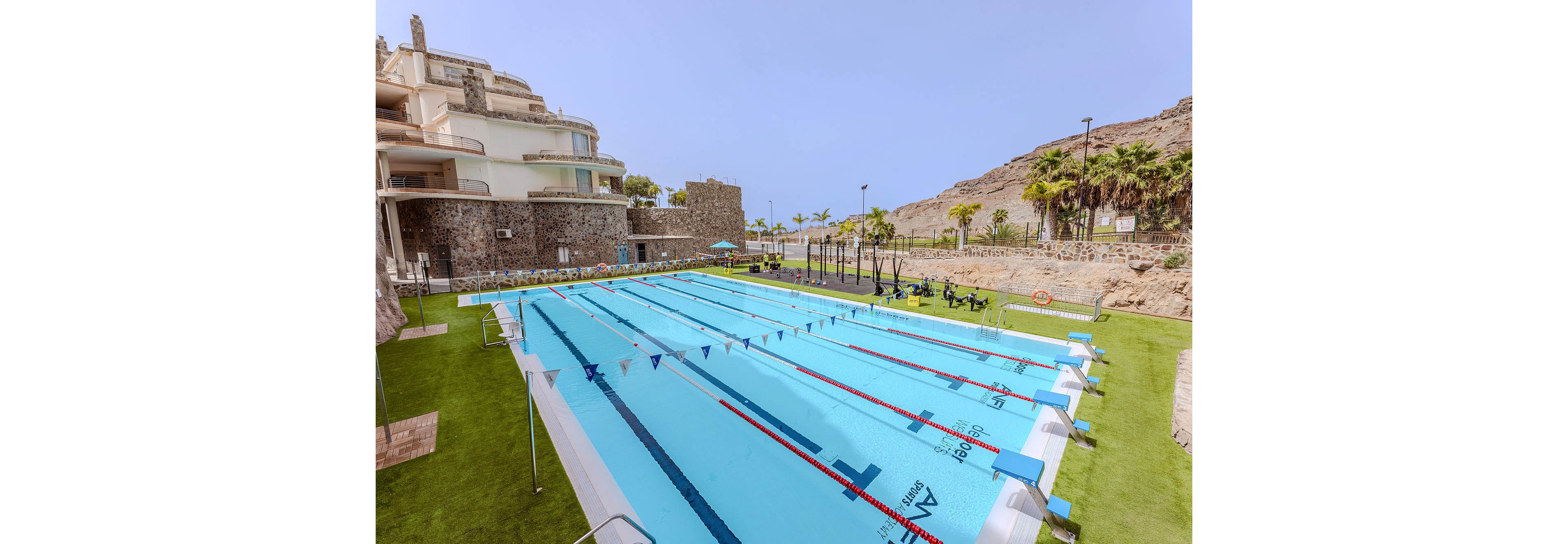 A large outdoor swimming pool is set against a backdrop of rocky hills and a modern resort building. The pool features multiple swim lanes, lane dividers, and starting blocks, with clear blue water and a grassy area surrounding it. Palm trees and lounge chairs line the poolside, creating a relaxed and sunny atmosphere. Visible numbers such as '2', '3', '4', and '5' are marked on the pool deck and starting blocks.