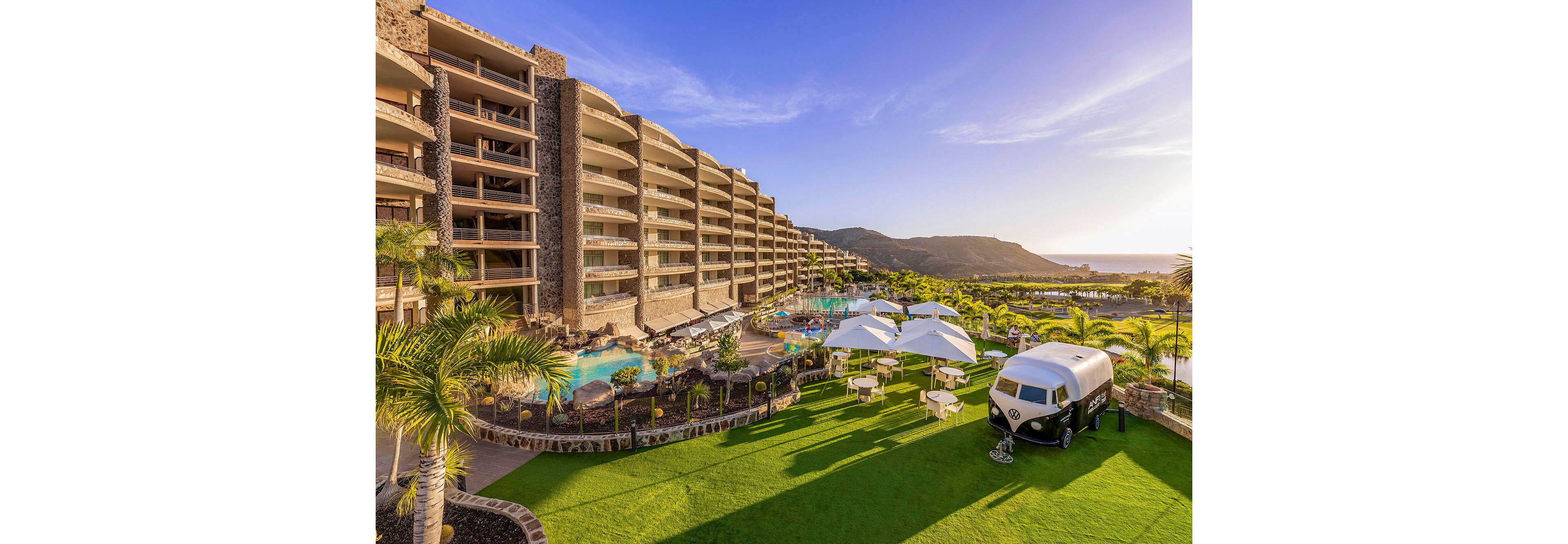 A modern multi-story resort overlooks a lush green lawn and a large swimming pool area, with palm trees and sun loungers. White umbrellas shade outdoor seating, and a vintage van is parked on the grass, adding a retro touch. The scene is bathed in warm sunset light, with mountains visible in the background and a clear blue sky overhead.