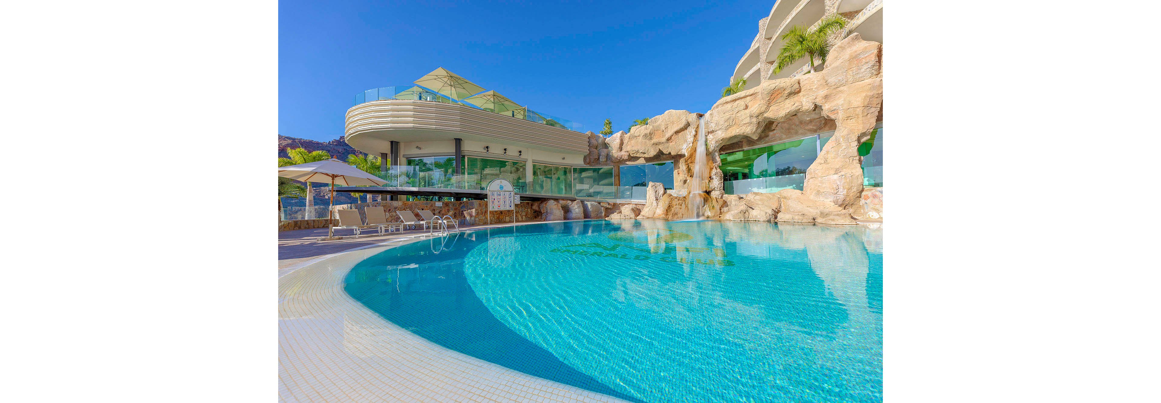 A luxurious outdoor swimming pool is the main focus, featuring a striking rock waterfall and clear blue water. The setting is a contemporary resort with curved architecture, sun loungers, and shaded seating areas. The sky is clear and blue, enhancing the bright, inviting atmosphere. No people are present, emphasizing relaxation and tranquility. The scene is visually dominated by natural stone textures and modern design elements.