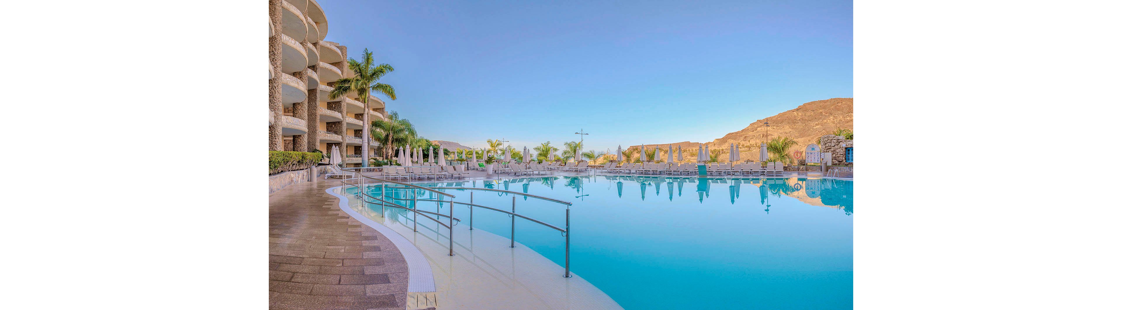 A large, tranquil swimming pool is the central focus of this luxury resort scene. The pool is surrounded by sun loungers, umbrellas, and tall palm trees, with modern curved hotel buildings on one side and a mountainous landscape in the background. The clear blue sky and calm water create a serene and inviting atmosphere, ideal for relaxation and leisure.