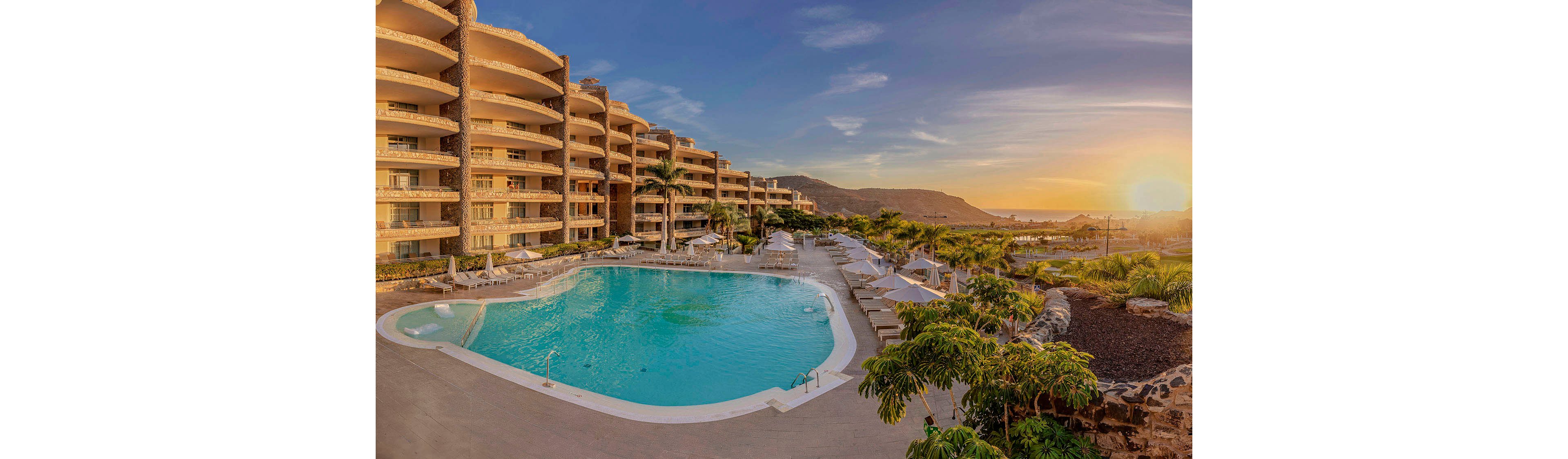 A large, inviting swimming pool is surrounded by sun loungers and umbrellas at a luxury resort. The scene is set during sunset, casting a warm golden glow over the curved multi-story hotel buildings and lush palm trees. In the background, mountains and landscaped gardens enhance the tranquil vacation atmosphere. The pool area is empty, emphasizing relaxation and exclusivity. No visible text or numbers are present in the image.