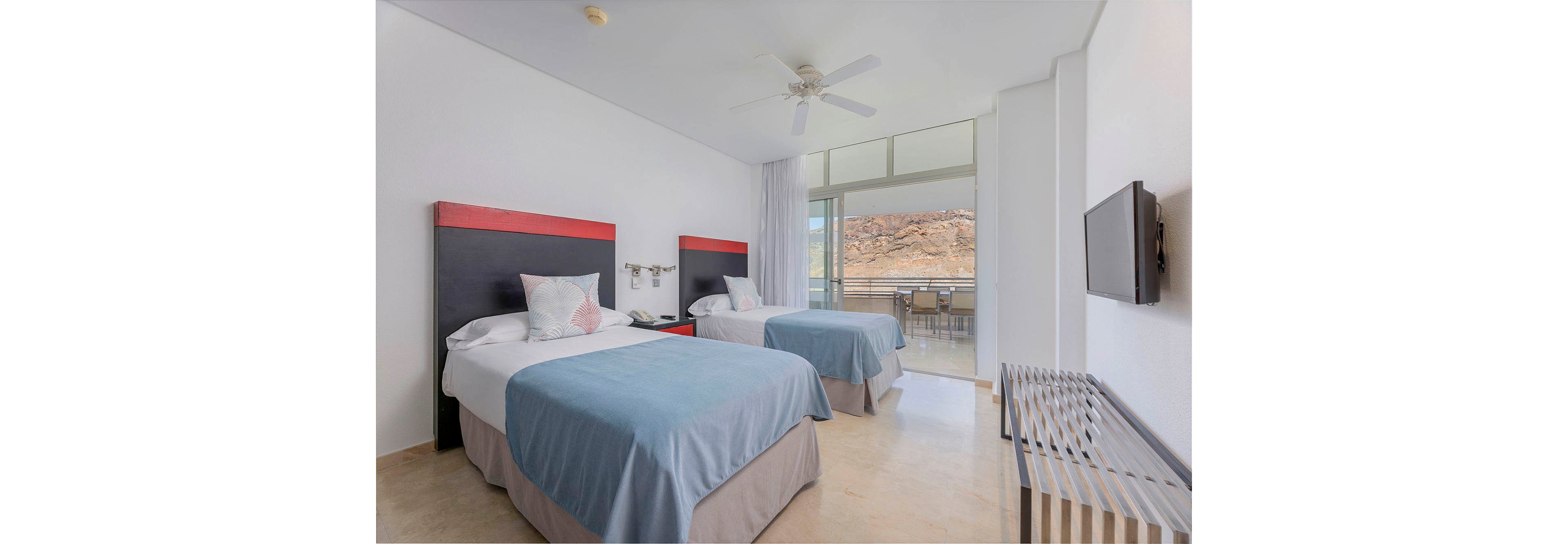 A bright hotel room features two twin beds with blue bedspreads and red-accented headboards. The space is decorated in a minimalist style with neutral tones and a ceiling fan overhead. Large windows open to a balcony with outdoor seating and a scenic mountain landscape visible in the background. The room includes a wall-mounted television and a luggage rack. No visible text or numbers are present in the image.