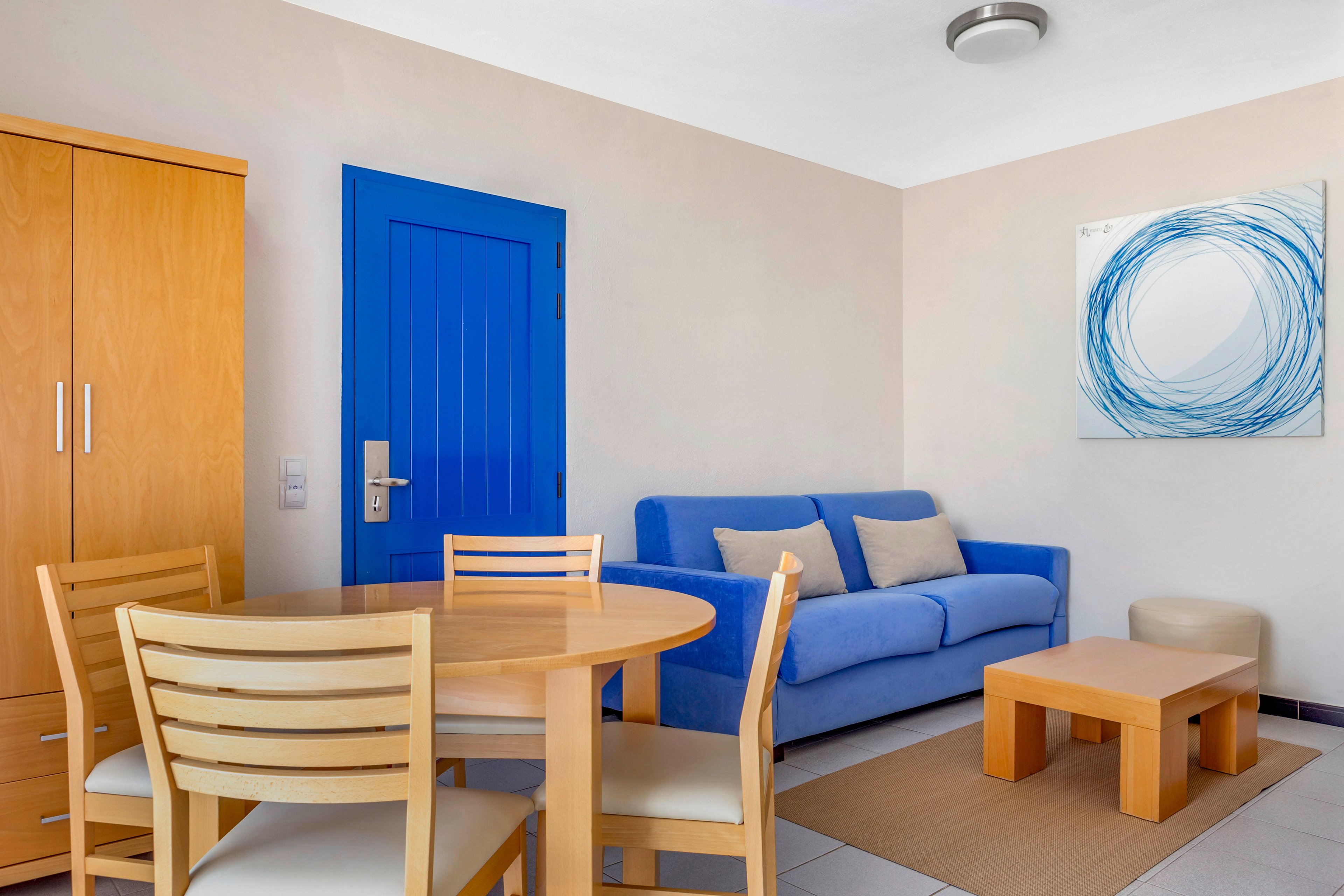 A contemporary living room features a bright blue sofa and matching blue door as focal points. Light wooden furniture, including a round dining table, chairs, and a low coffee table, complement the neutral walls and tiled floor. A minimalist abstract artwork with blue circular lines hangs above the sofa, enhancing the cohesive color palette. The space appears clean, organized, and inviting, with soft cushions adding comfort to the seating area.