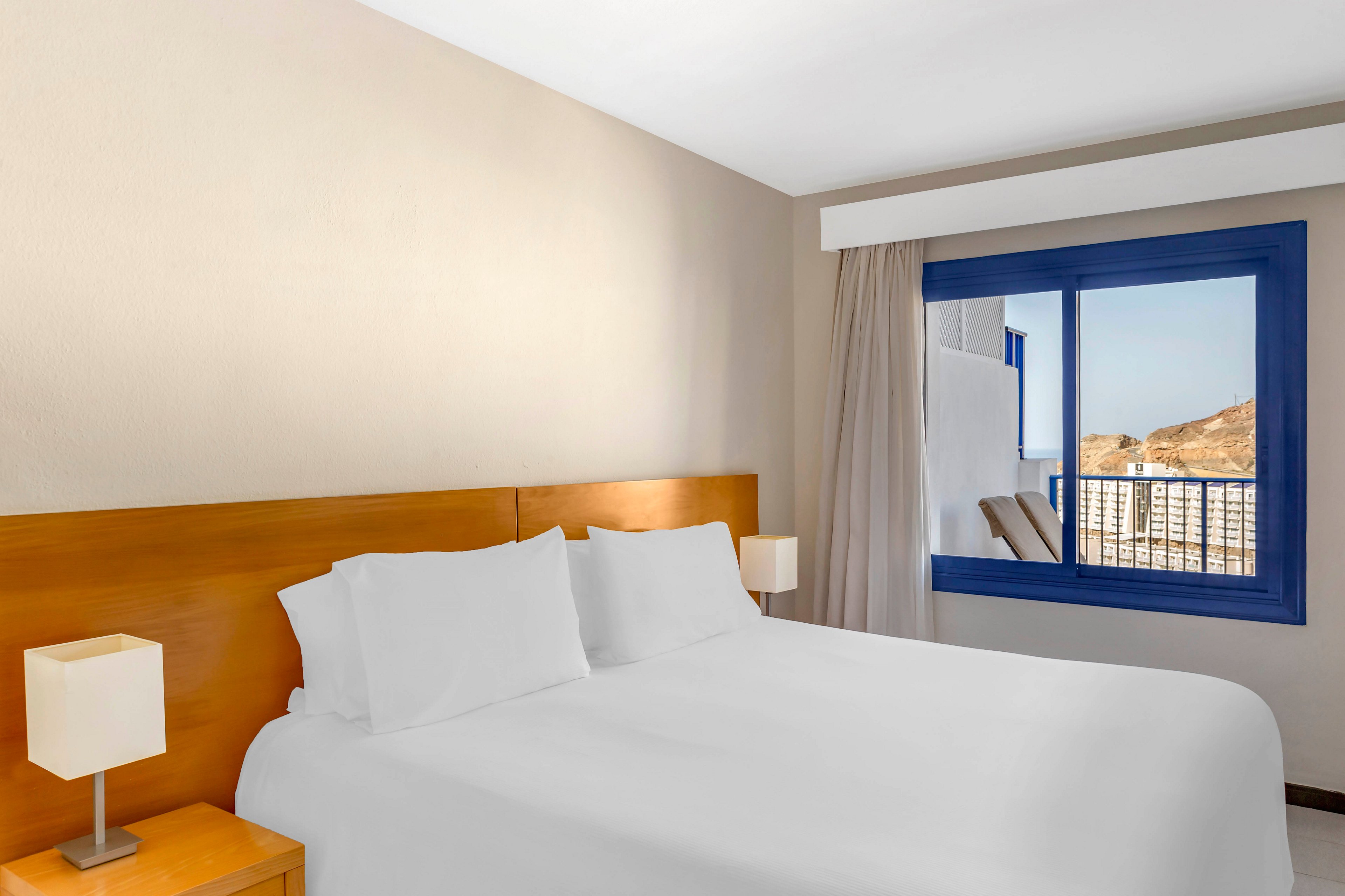A neatly made double bed with white linens is the focal point of this bright hotel room. Wooden furniture and matching bedside lamps add warmth to the minimalist decor. A large window with blue trim opens onto a balcony, where two chairs face a distant hillside and buildings. Natural daylight fills the room, creating a clean and inviting atmosphere.