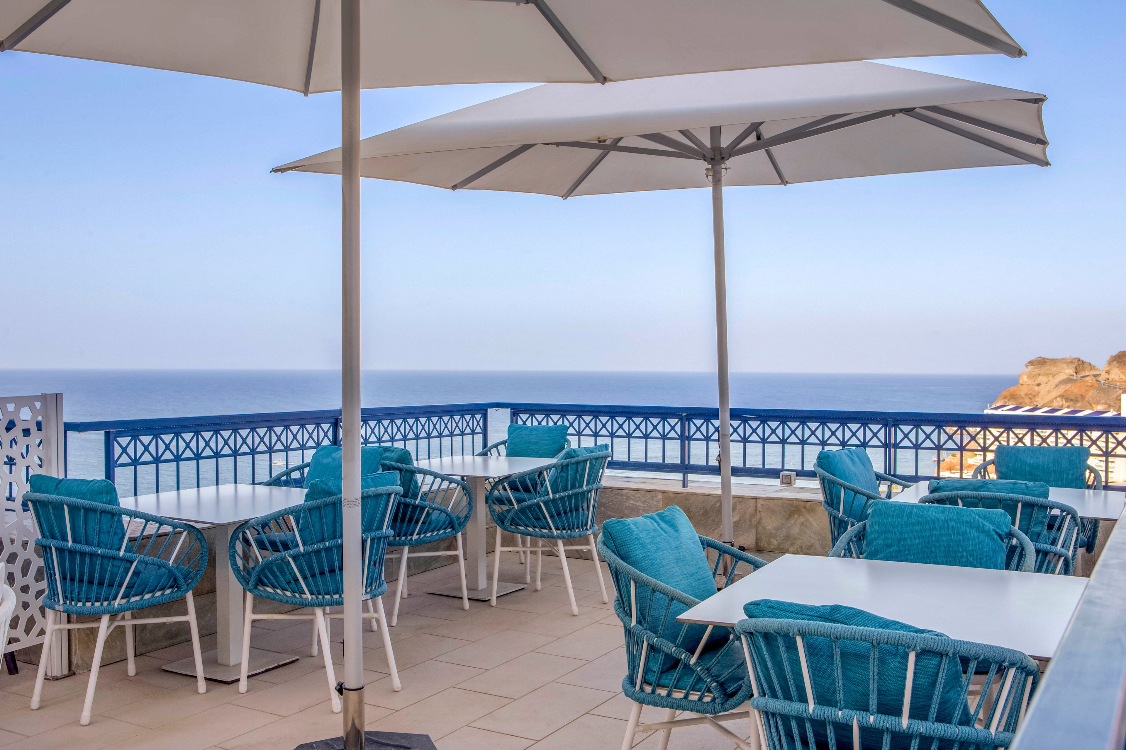 An outdoor terrace cafe overlooks a calm ocean under a clear blue sky. White tables and teal cushioned chairs are arranged neatly beneath large white umbrellas. A blue railing and decorative white panel frame the seating area, enhancing the coastal atmosphere. The scene is quiet and inviting, suggesting a relaxed seaside dining experience.