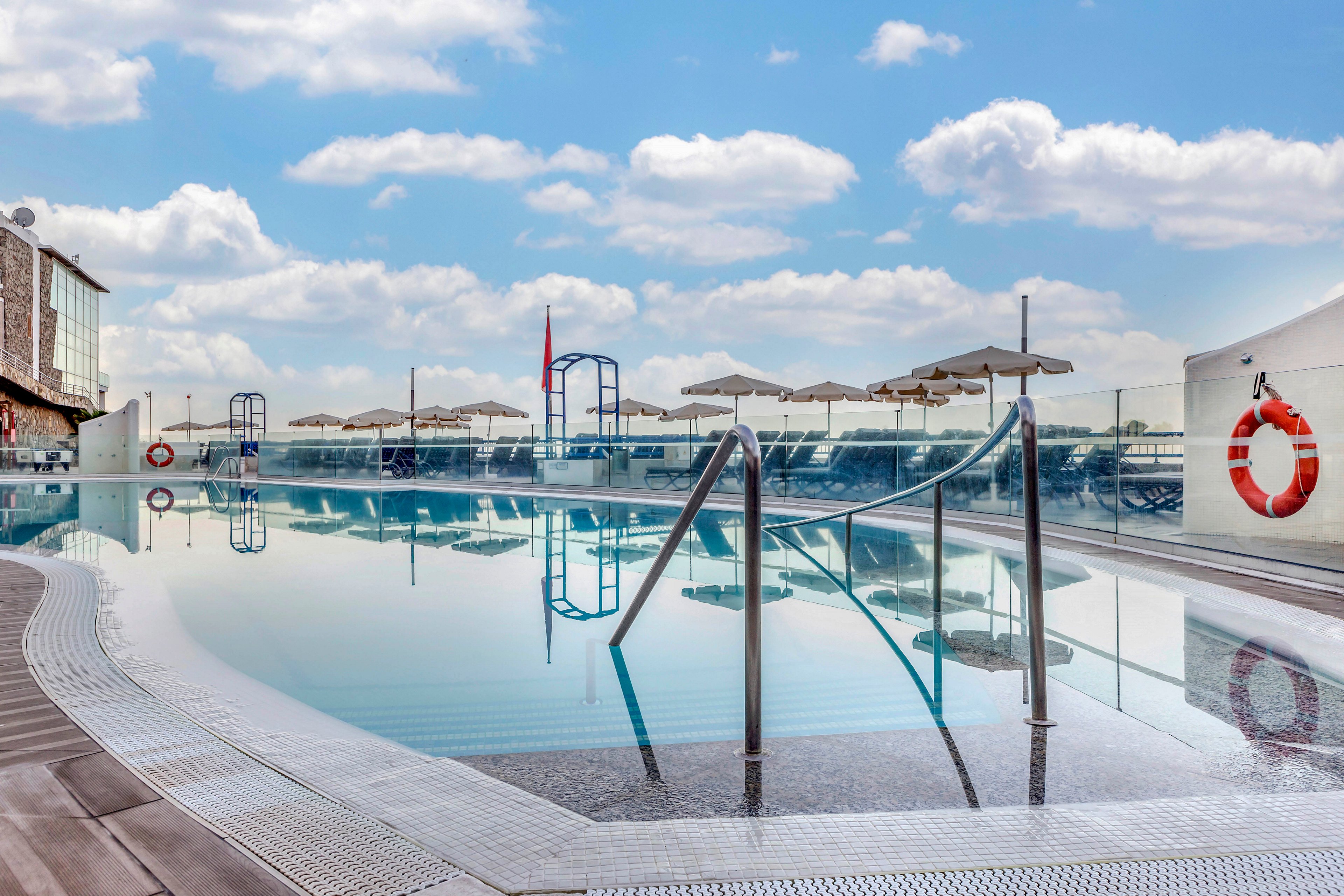 A calm rooftop swimming pool reflects a bright blue sky with scattered clouds. Metal handrails lead into the still water, and sun umbrellas line the far side of the pool. A lifebuoy hangs on the glass barrier, emphasizing safety around the water. The modern urban setting suggests a hotel or residential terrace designed for relaxation and leisure.