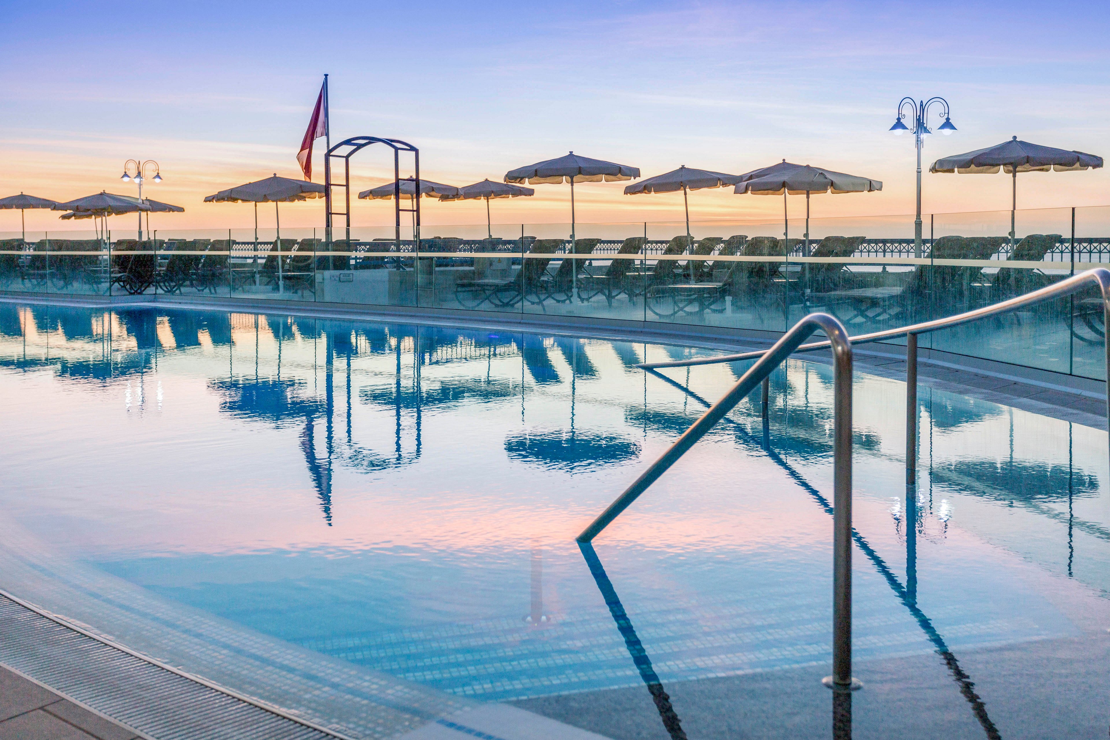 A calm outdoor swimming pool reflects the pastel colors of the early morning sky. Sun loungers and closed umbrellas line the far side of the pool, facing the ocean horizon. Metal handrails lead into the still water, emphasizing a tranquil and inviting atmosphere. Soft sunrise light and gentle reflections create a peaceful resort setting by the sea.