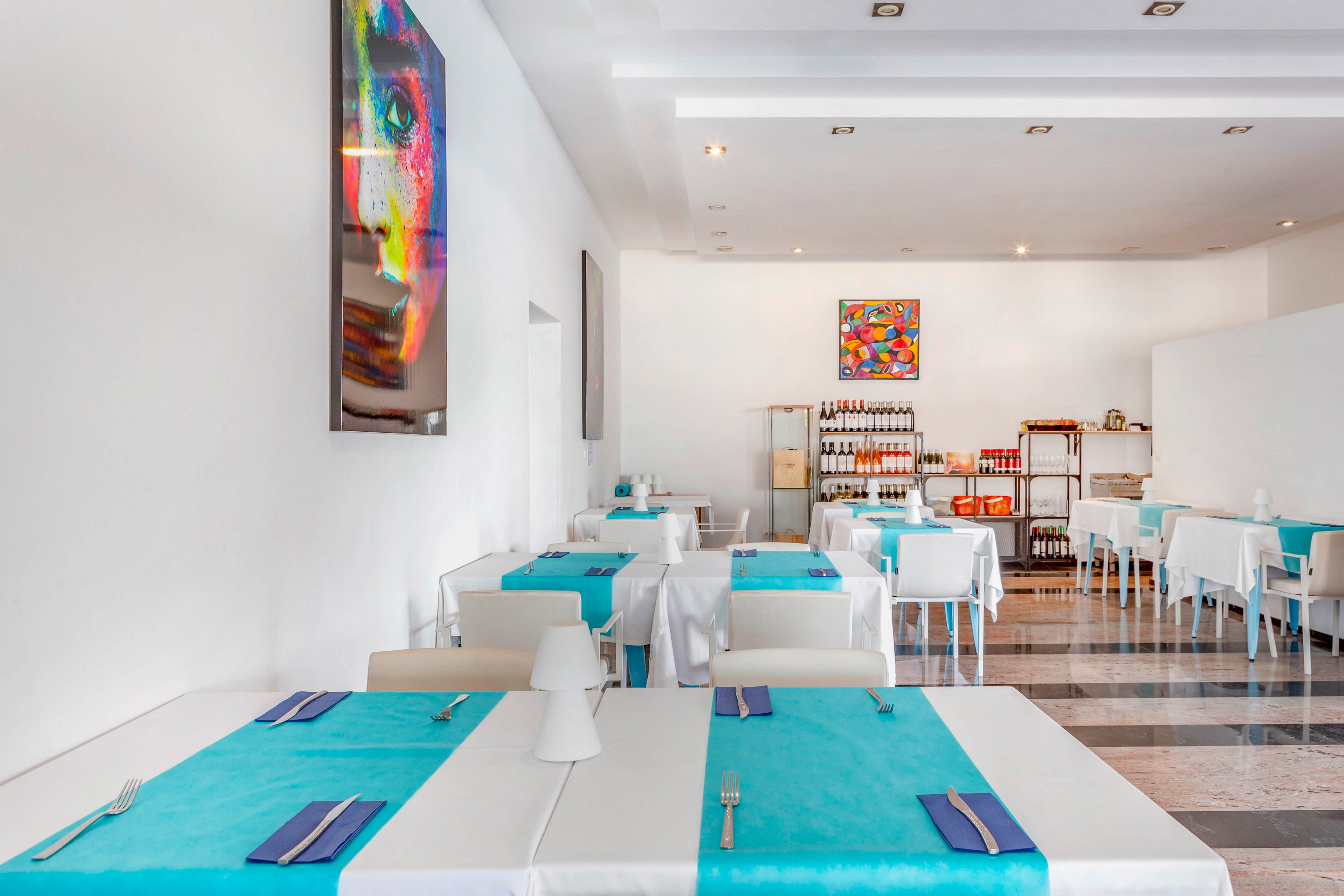 A bright, modern restaurant interior features neatly arranged tables covered with white cloths and turquoise runners. The space is minimalist, with white walls, white chairs, and clean lines creating an airy atmosphere. Colorful abstract artwork hangs on the walls, adding vibrant accents to the otherwise neutral setting. Shelves at the back display bottles and jars, suggesting a well-stocked dining area ready for guests.