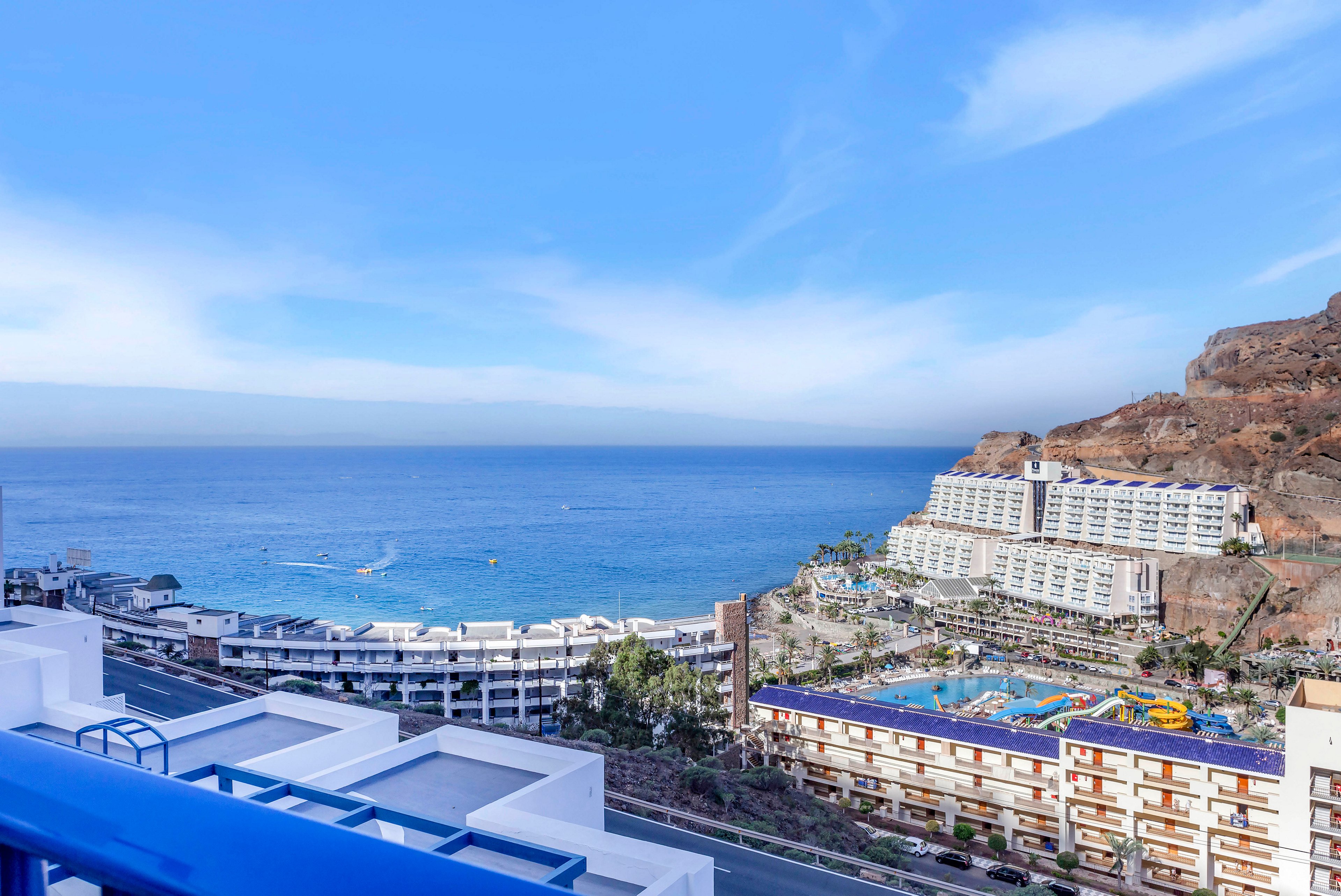 A coastal resort town stretches along a bright blue ocean under a clear sky. Multiple terraced hotel buildings and apartments line the waterfront, with a large outdoor swimming pool complex in the foreground. The scene includes a winding coastal road, palm trees, and rocky hills rising behind the resorts. Small boats can be seen on the calm sea, emphasizing a relaxed vacation atmosphere.