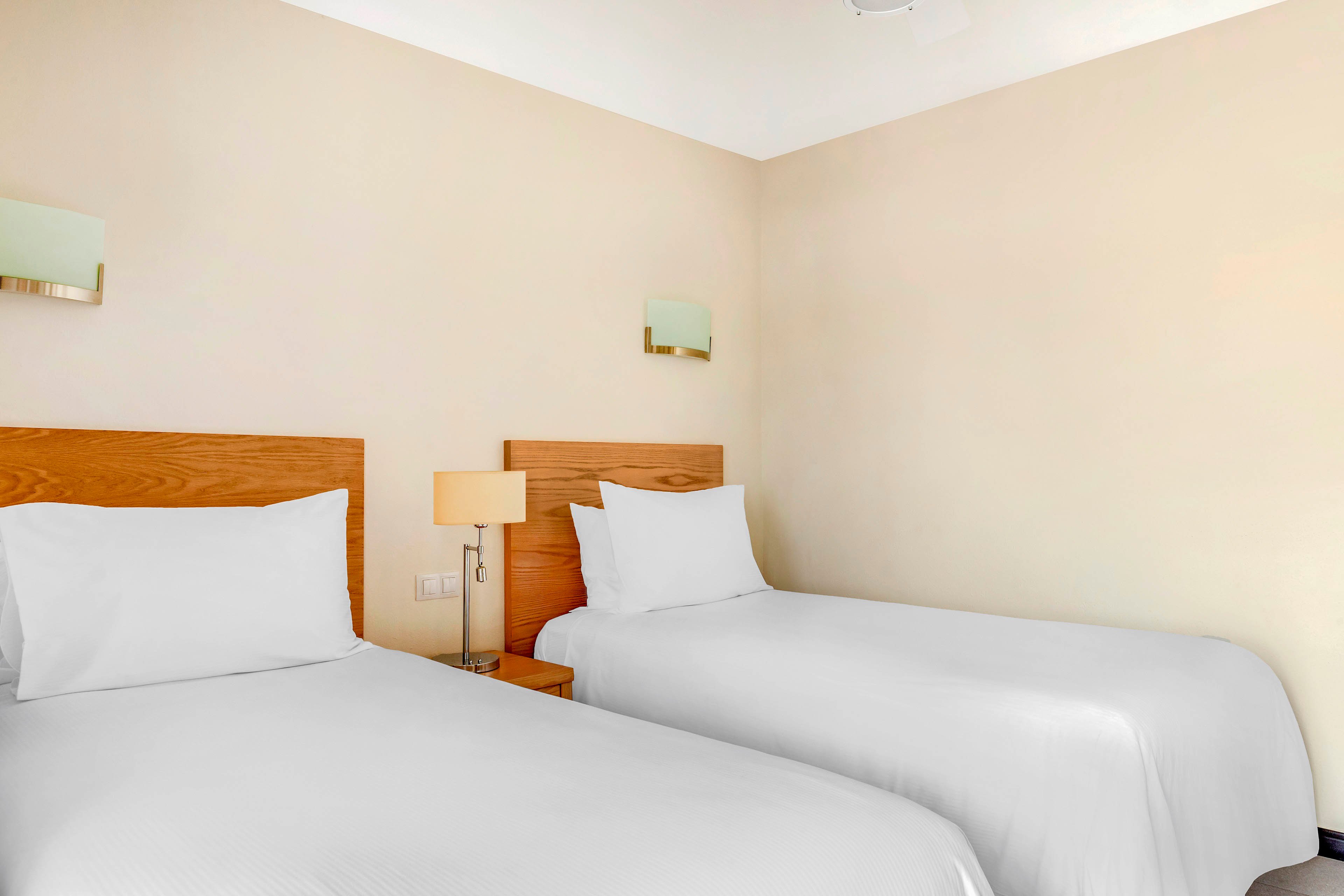 A simple hotel room features two neatly made twin beds with white linens and wooden headboards. A small bedside table with a lamp sits between the beds, adding a warm accent to the neutral decor. Soft wall-mounted lights and a beige wall create a calm, minimalist atmosphere. The overall scene suggests a clean, comfortable, and functional guest room.