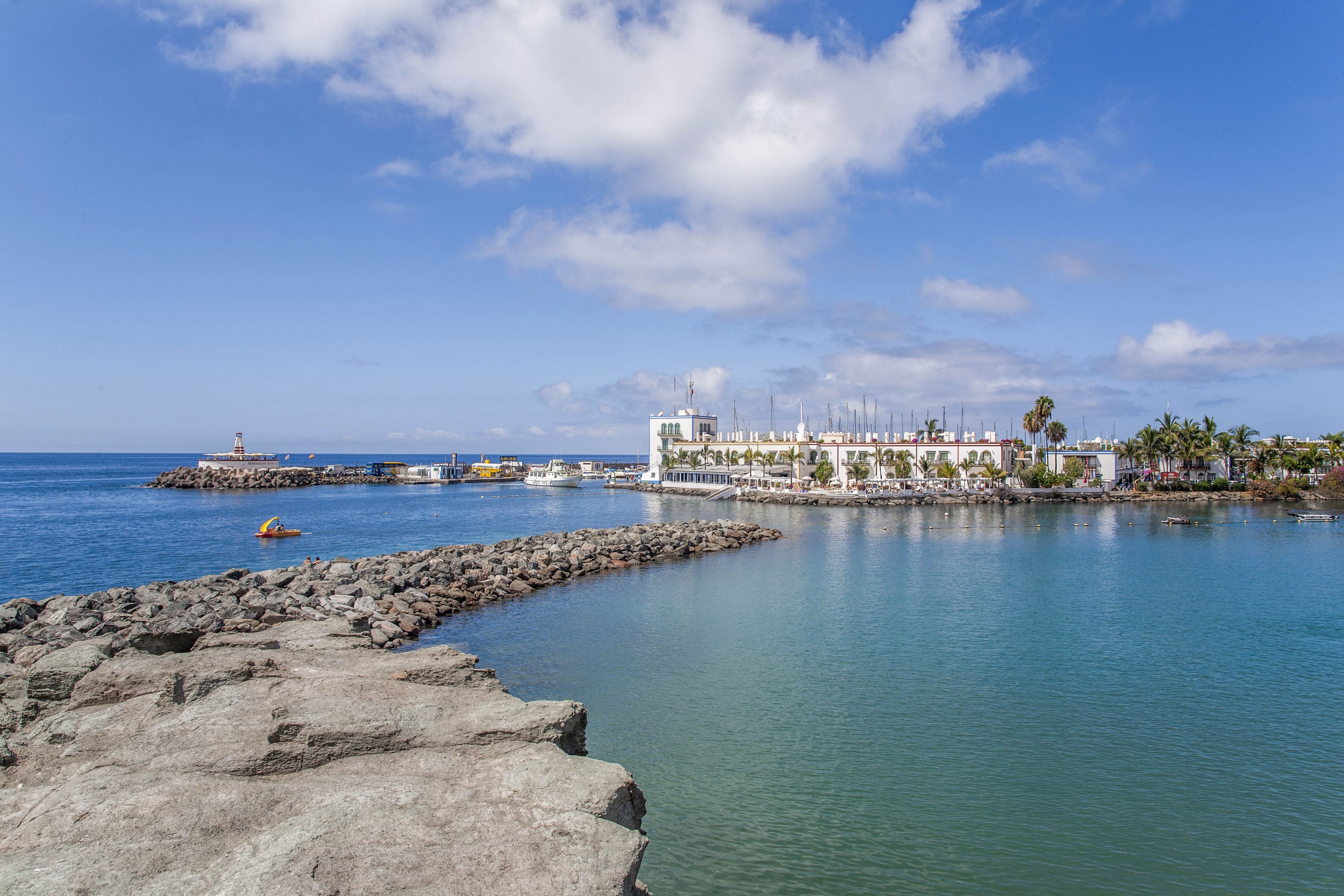 A calm coastal marina is viewed from a rocky breakwater extending into the sea. The harbor area features white buildings, moored boats, and a small lighthouse near the entrance. Clear blue water fills the foreground, reflecting the sky and scattered clouds above. Palm trees and waterfront structures line the distant shore, creating a relaxed seaside atmosphere.