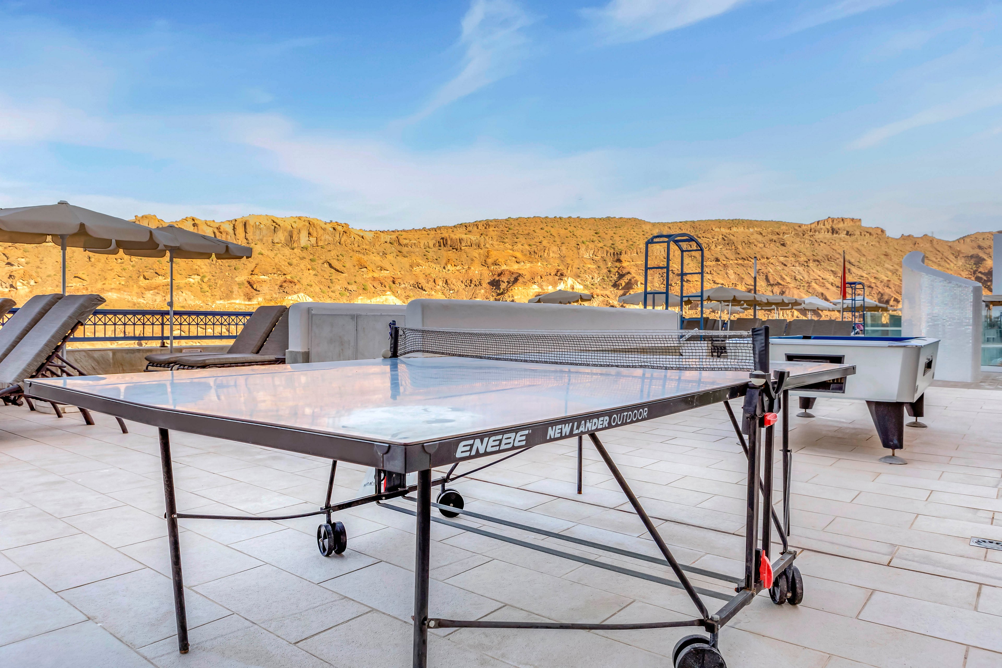 An outdoor ping pong table stands on a spacious tiled terrace under a clear blue sky. Lounge chairs and umbrellas line the patio, suggesting a relaxing recreational area. Rocky hills and a distant landscape form the backdrop beyond the terrace wall. The scene feels bright and open, emphasizing leisure and outdoor activity.