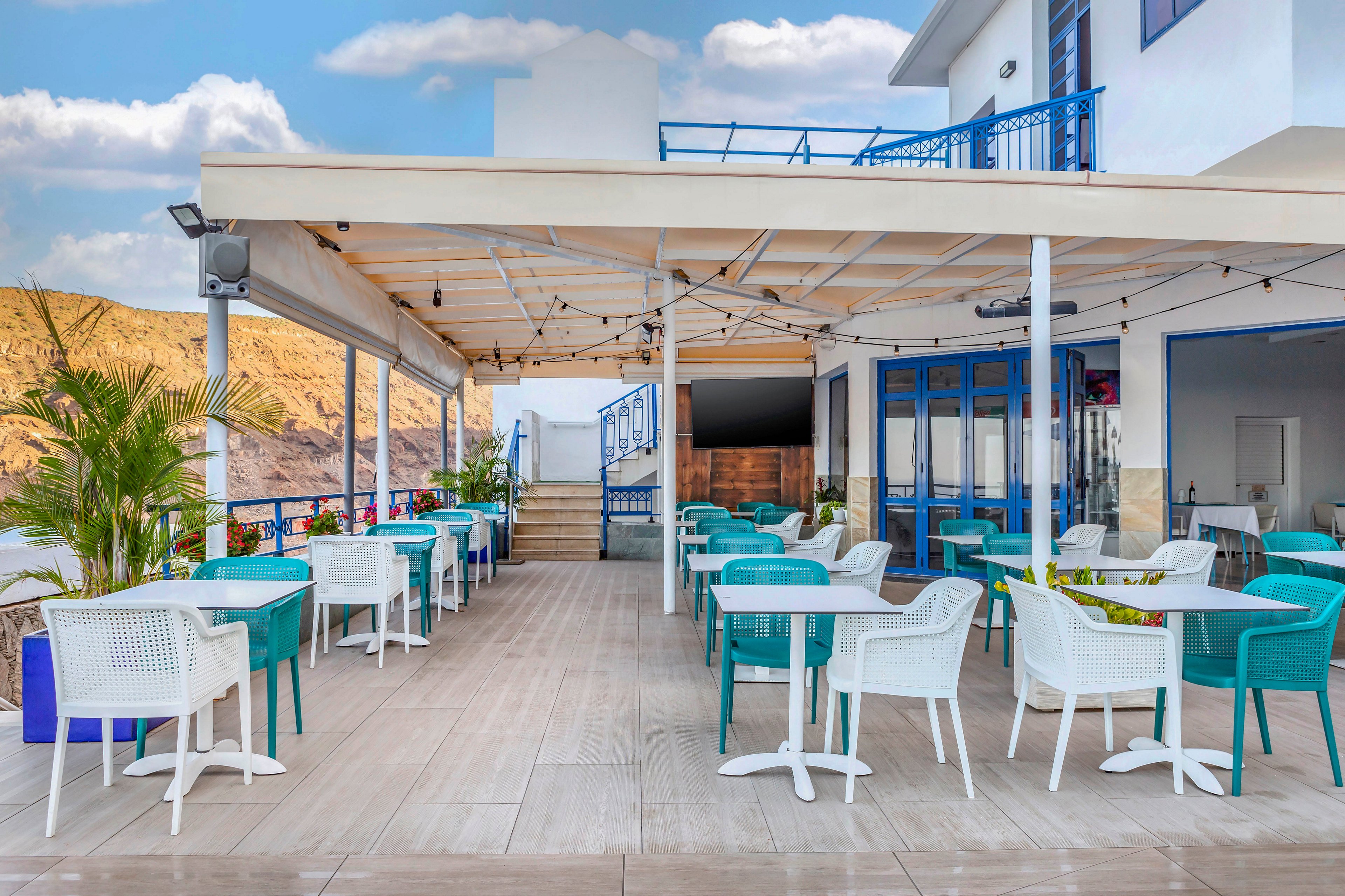 An open-air café terrace features rows of white and turquoise chairs around matching tables. The setting overlooks a coastal landscape with hills in the background under a partly cloudy sky. A light wooden pergola with string lights covers the seating area, creating a relaxed atmosphere. Blue railings, doors, and accents on the building enhance the bright, coastal aesthetic.