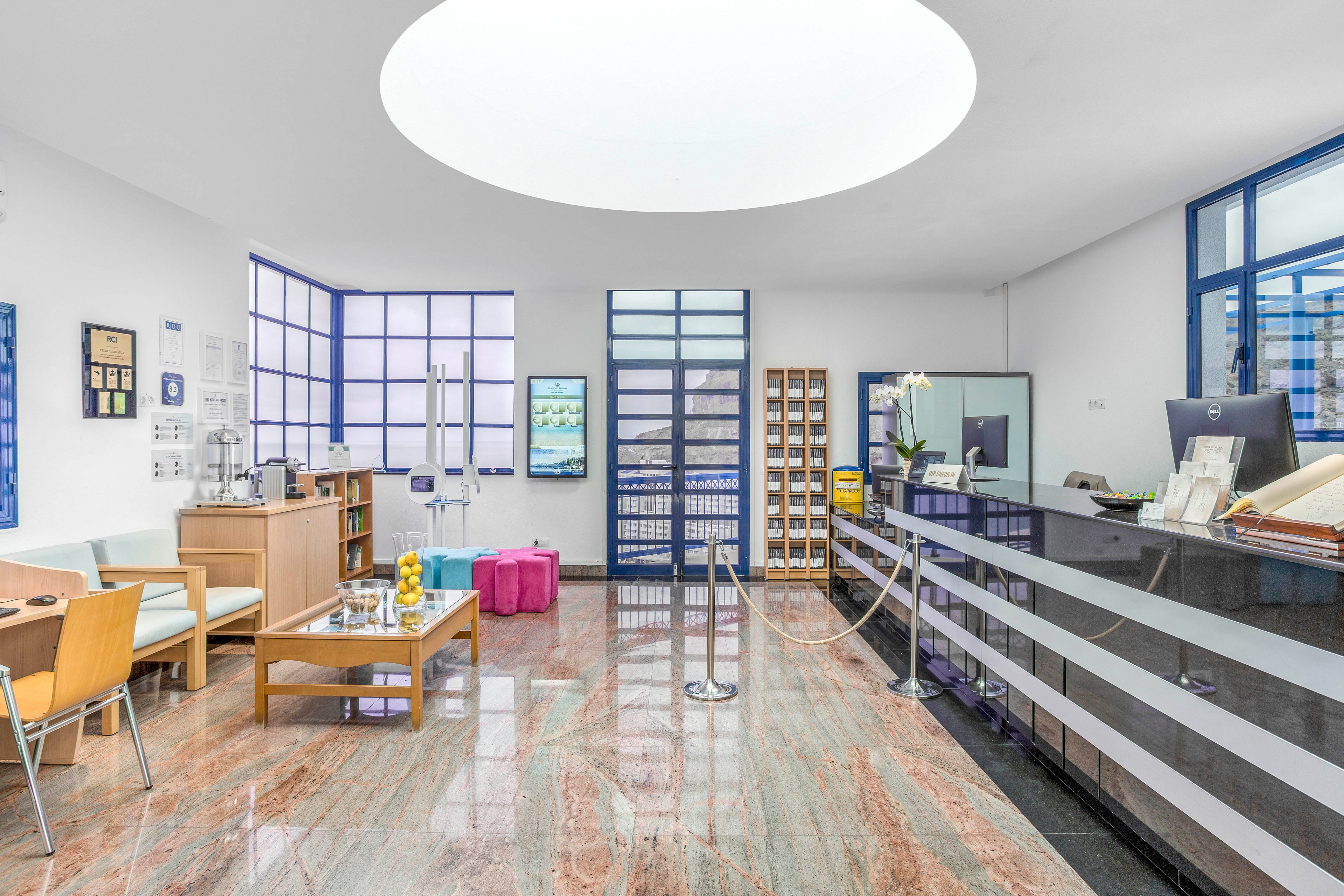 A contemporary reception area features a sleek front desk and polished marble floor under a large circular skylight. The space includes a seating area with wooden tables, colorful cube stools, and chairs arranged for visitors. Blue-framed windows and doors bring in natural light, while shelves and display units line the walls. A rope barrier guides guests toward the reception counter, creating an orderly and welcoming atmosphere.