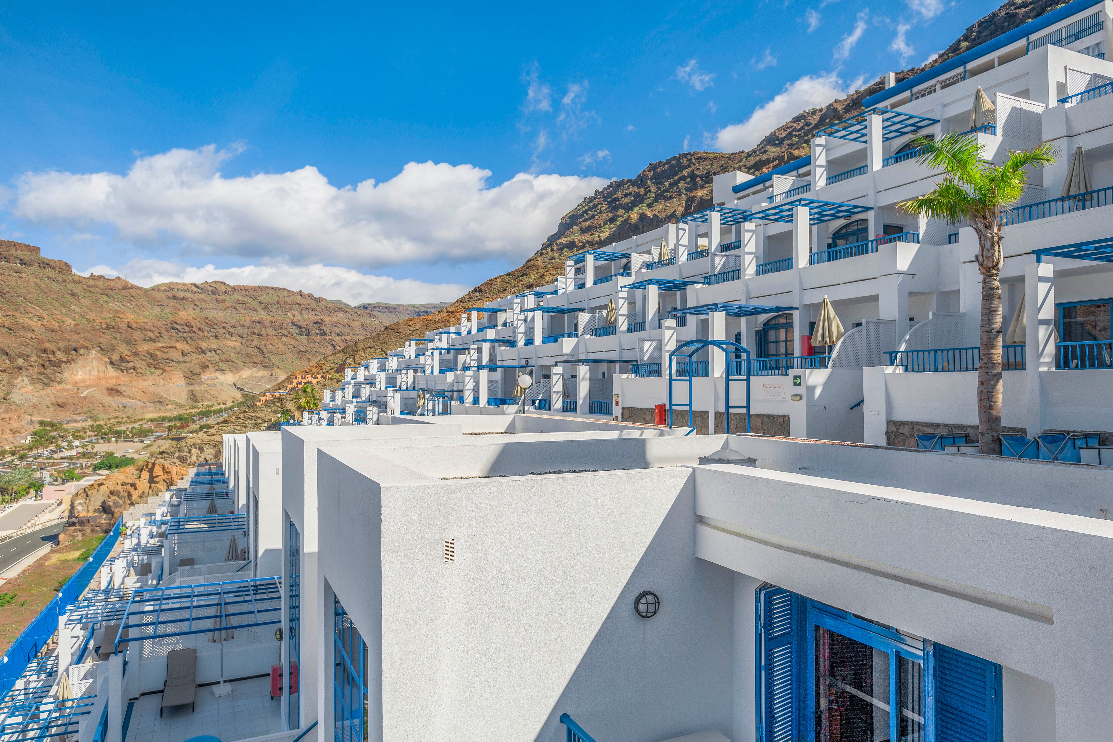 A series of white terraced resort buildings steps up a rocky hillside under a bright blue sky. Each unit features blue railings, balconies, and windows that create a striking contrast against the white walls. The setting appears to be a sunny coastal or mountain destination with clear weather and scattered clouds. The overall scene conveys a clean, modern Mediterranean or island-style architecture with strong geometric lines.