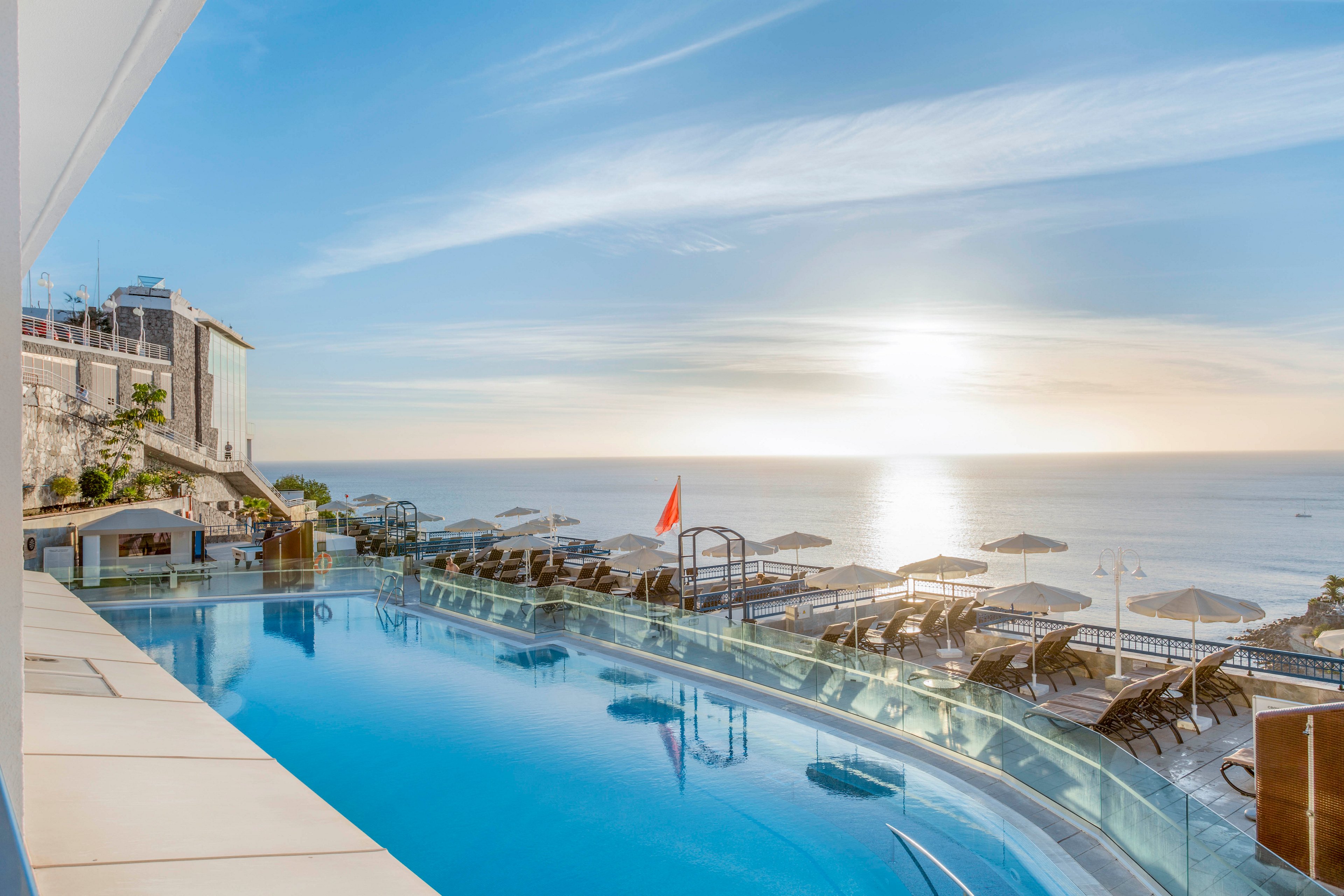 A serene infinity pool reflects the soft light of a setting sun over the ocean. Lounge chairs and umbrellas line the poolside terrace, suggesting a luxurious seaside resort. Modern buildings rise along the left side, framing the expansive coastal view. The calm water, clear sky, and warm sunlight create a tranquil vacation atmosphere.