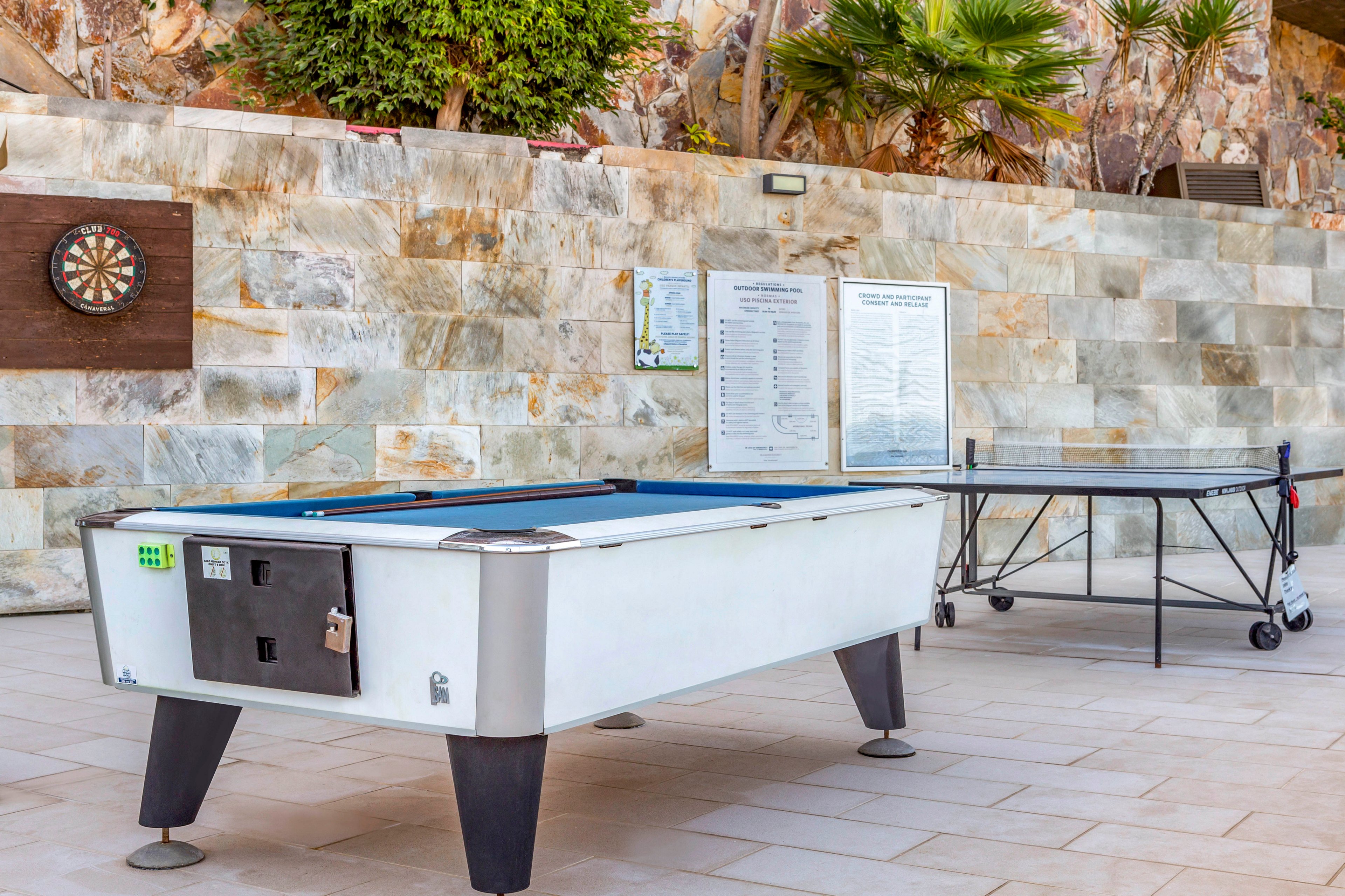 An outdoor leisure area features a white pool table as the main focus. Behind it, a ping pong table is set up against a stone wall. A dartboard and framed information boards are mounted on the wall, suggesting a recreational or resort setting. The scene is bright and clean, with tiled flooring and greenery above the wall adding a relaxed atmosphere.