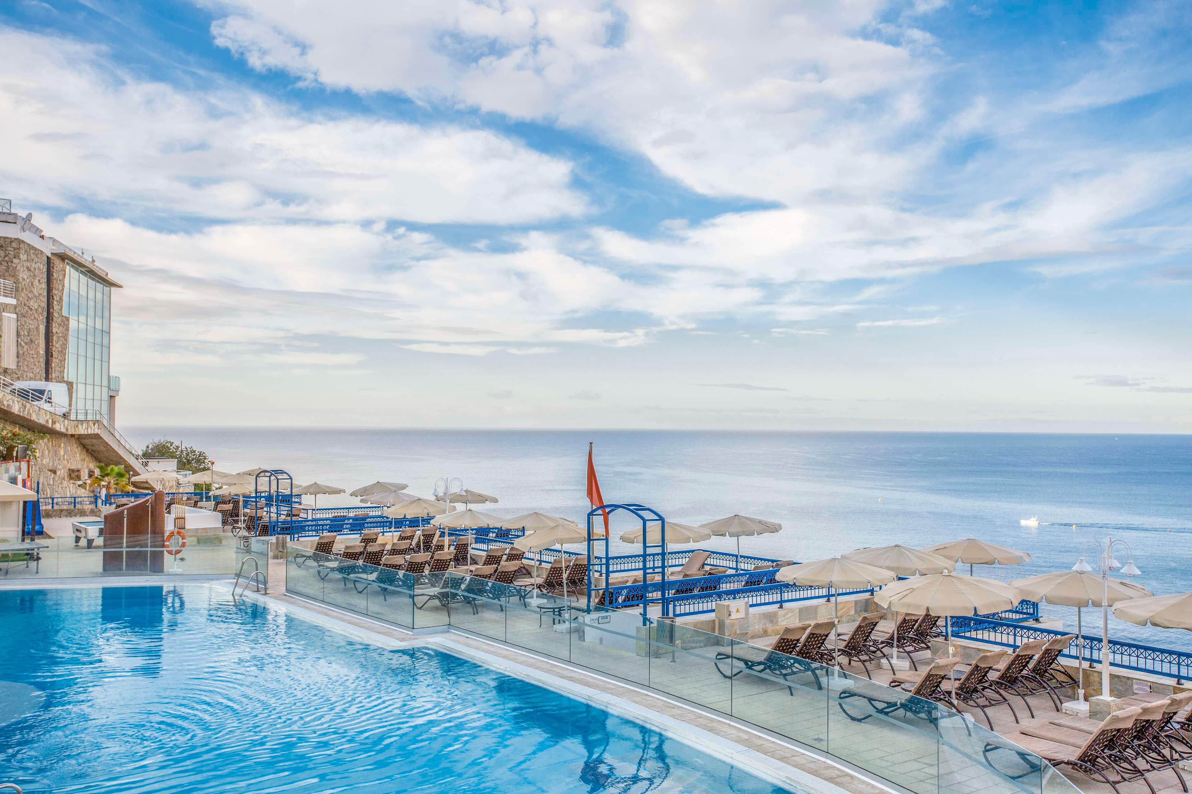 A large infinity pool overlooks a calm, expansive ocean at a seaside resort. Numerous lounge chairs and umbrellas line the pool deck, facing the water for relaxation and sunbathing. Glass railings separate the pool area from the sea view, enhancing the open, airy atmosphere. The sky is partly cloudy with soft light, creating a tranquil and inviting coastal scene.