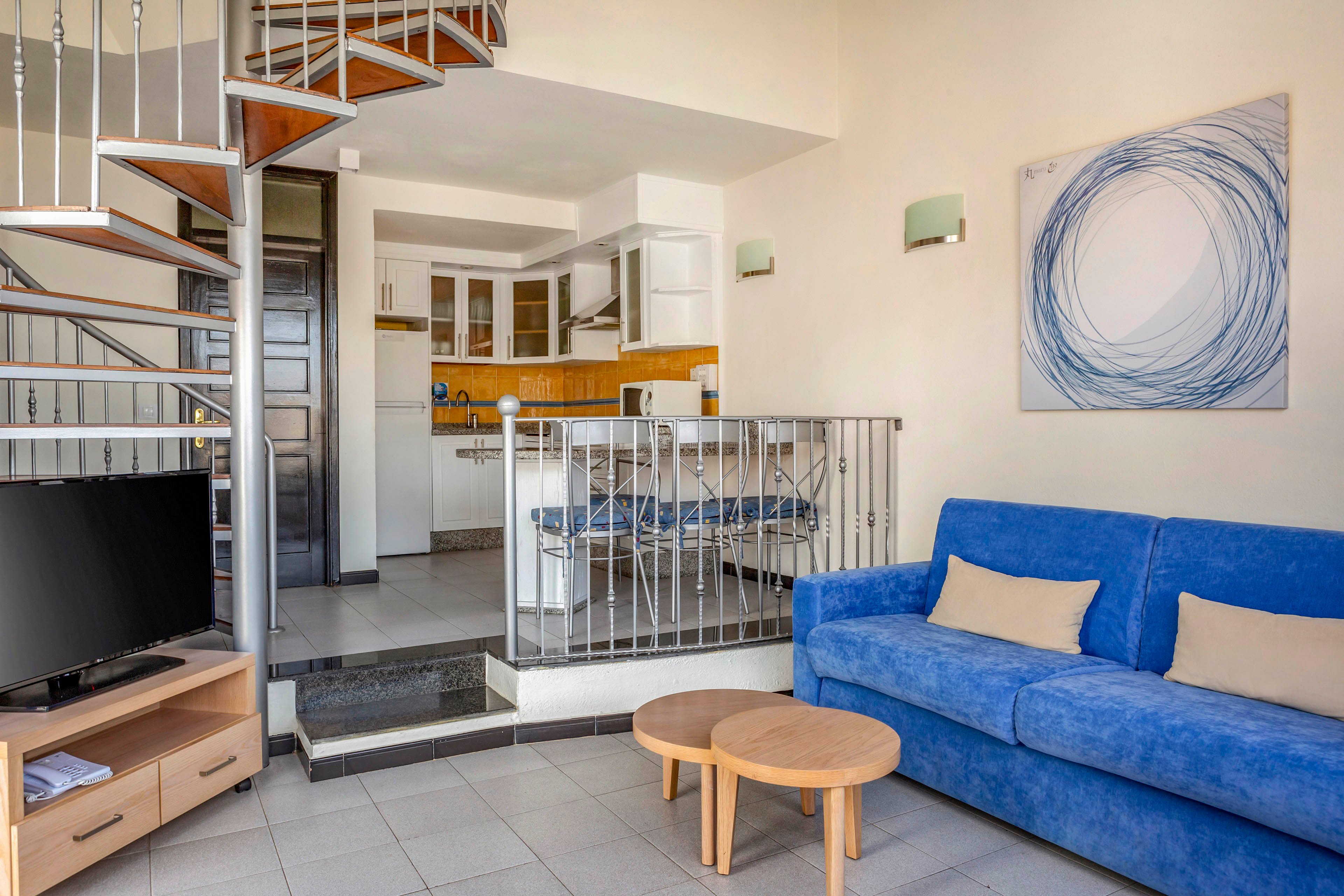 A bright modern living room features a blue sofa with beige cushions and a small round wooden coffee table. A metal spiral staircase leads up from the corner, adding a loft-like feel to the space. In the background, an open kitchen with white cabinets and yellow accents is visible behind a metal railing. A flat-screen television sits on a low wooden stand, and abstract wall art decorates the light-colored walls.