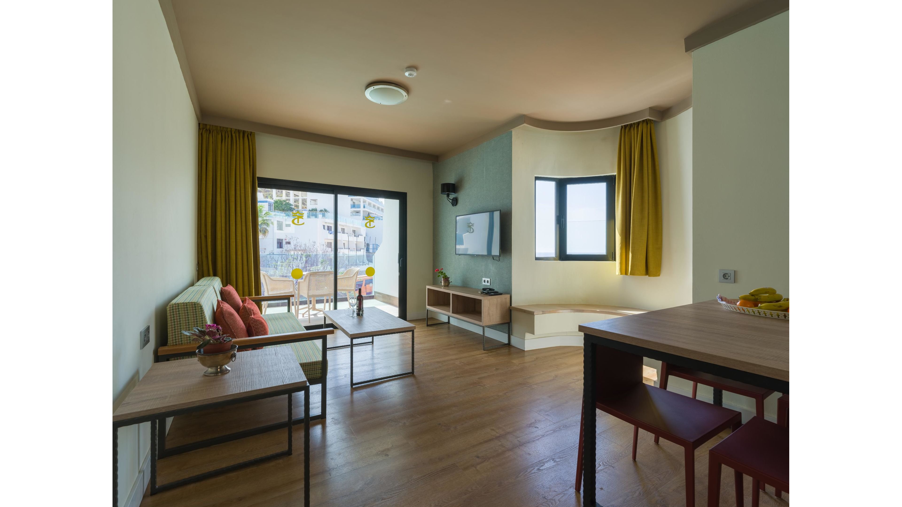 A contemporary living room features a sofa with orange cushions and simple wooden tables on a warm wood floor. Large glass doors open to a balcony, revealing a view of white buildings outside. A wall-mounted television and minimalist media console sit opposite the seating area, with a small decorative plant adding color. Yellow curtains and soft natural light create an inviting, relaxed atmosphere.
