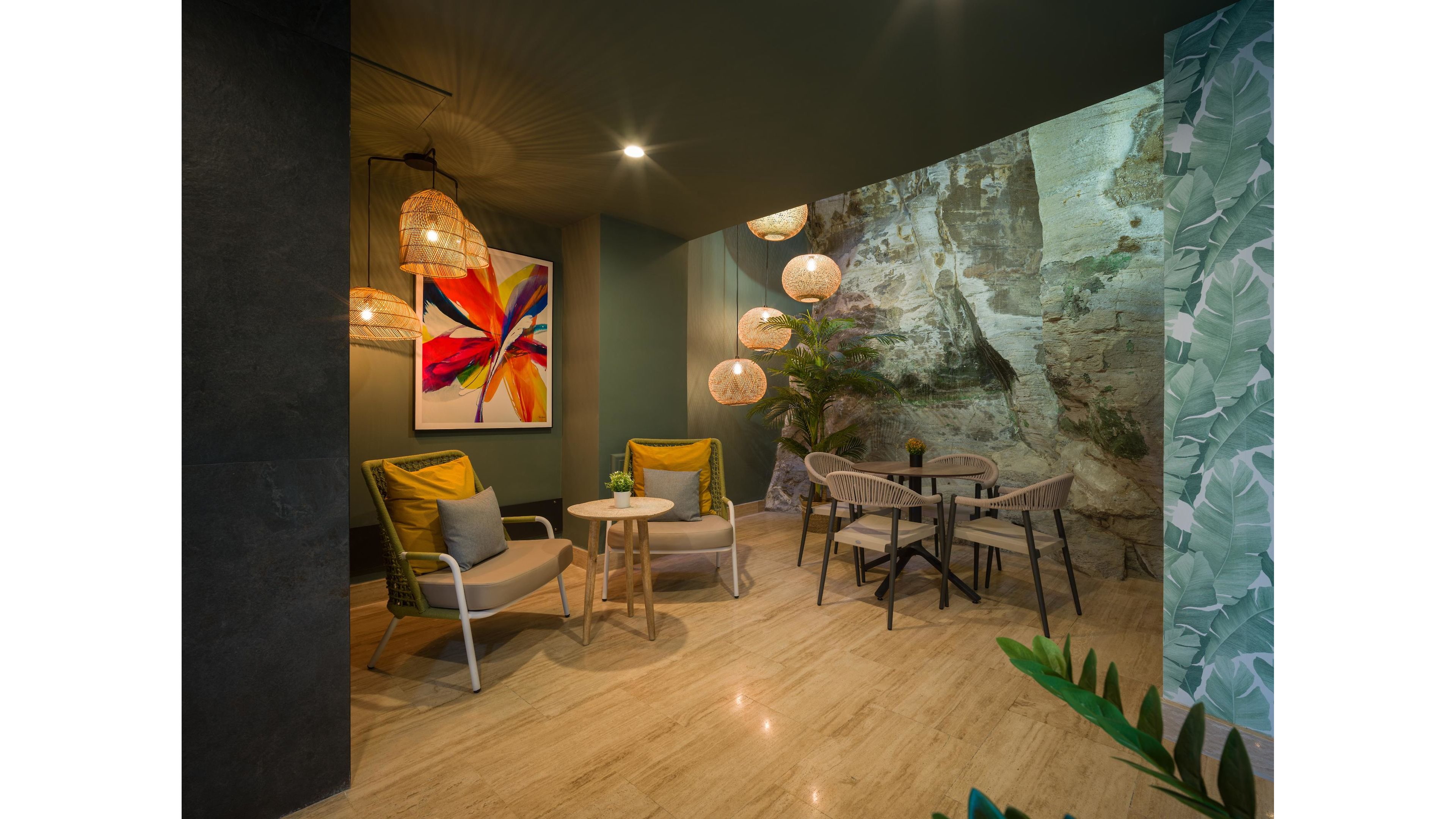 A cozy indoor lounge area features a small seating arrangement with cushioned armchairs and a round wooden table. Nearby, a second table with minimalist chairs sits under hanging woven pendant lights. The space is decorated with a large colorful abstract flower painting and a wall mural resembling a rocky cliff. Green plants and patterned wallpaper add a tropical touch, while polished flooring reflects the warm ambient lighting.