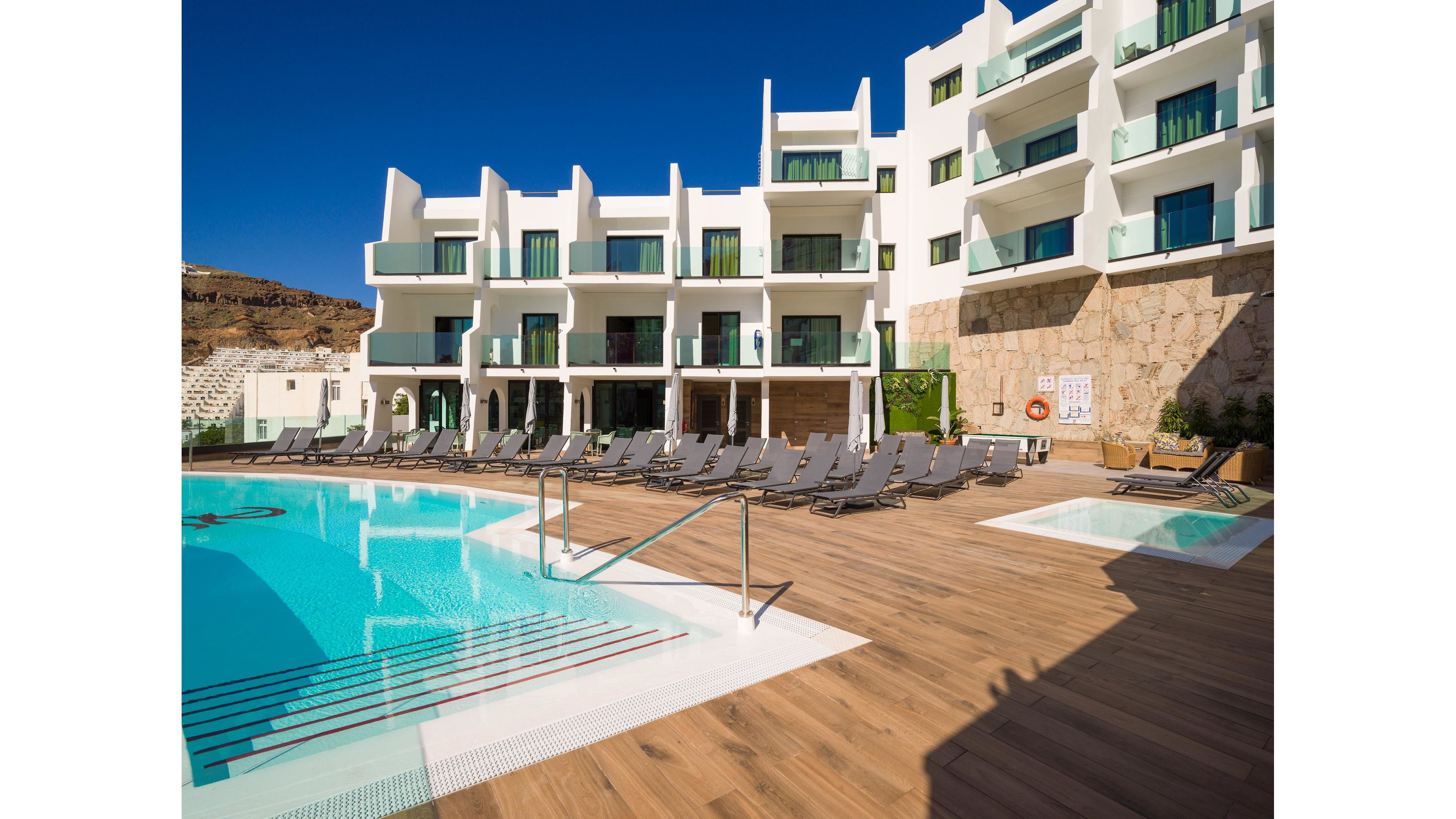 A contemporary white hotel building overlooks a clear blue outdoor swimming pool. Numerous gray sun loungers line the wooden deck, facing the water under a bright sunny sky. Glass balcony railings and large windows give the architecture a modern, minimalist feel. Rocky hills rise in the background, enhancing the vacation resort atmosphere.
