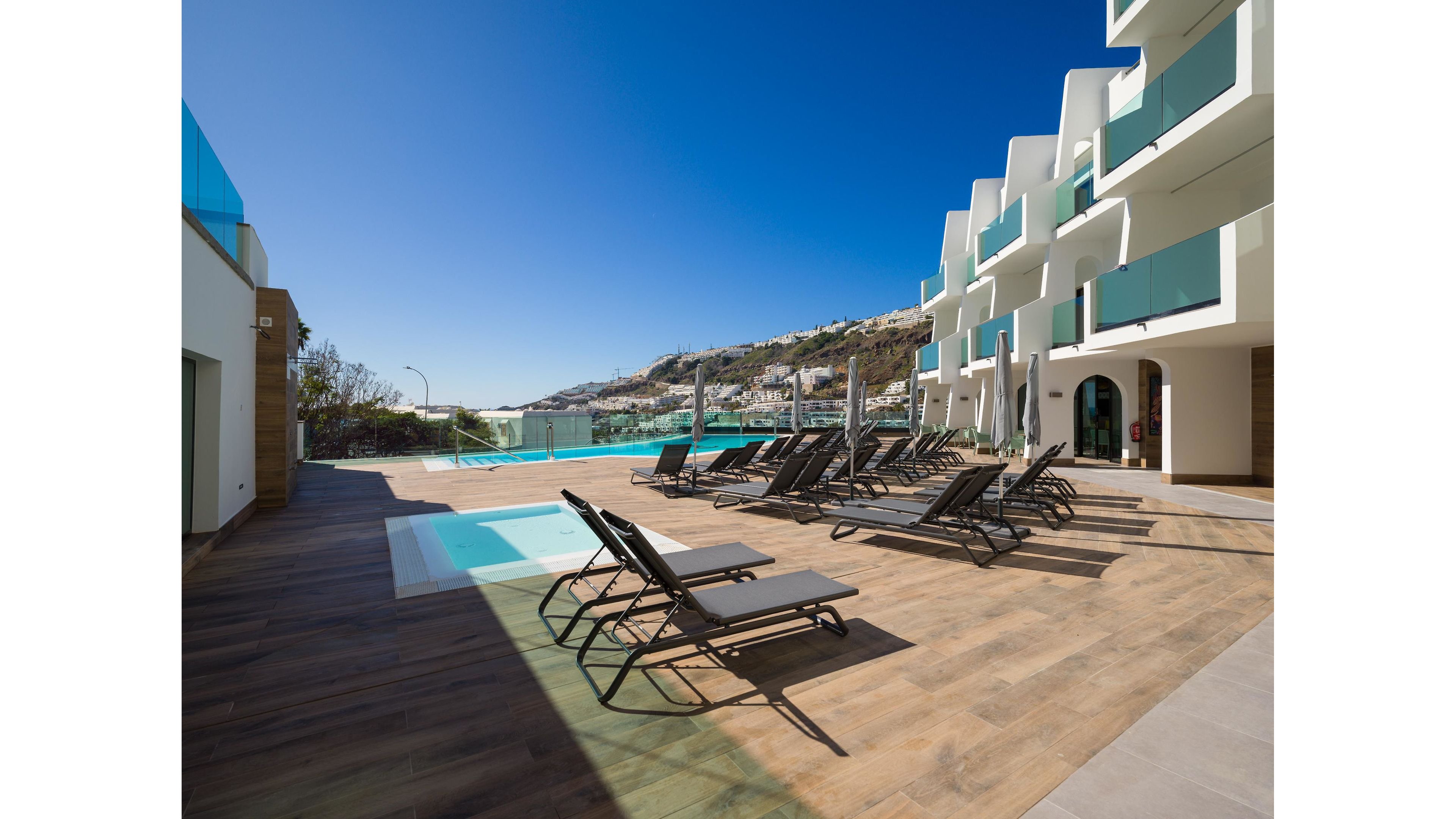 A modern hotel terrace features a row of dark lounge chairs beside a sleek outdoor swimming pool. The scene overlooks a hillside town under a clear blue sky, suggesting a warm, sunny day. Glass railings and contemporary white architecture frame the pool area, enhancing the minimalist design. The empty loungers and calm water create a tranquil, resort-like atmosphere ideal for relaxation.