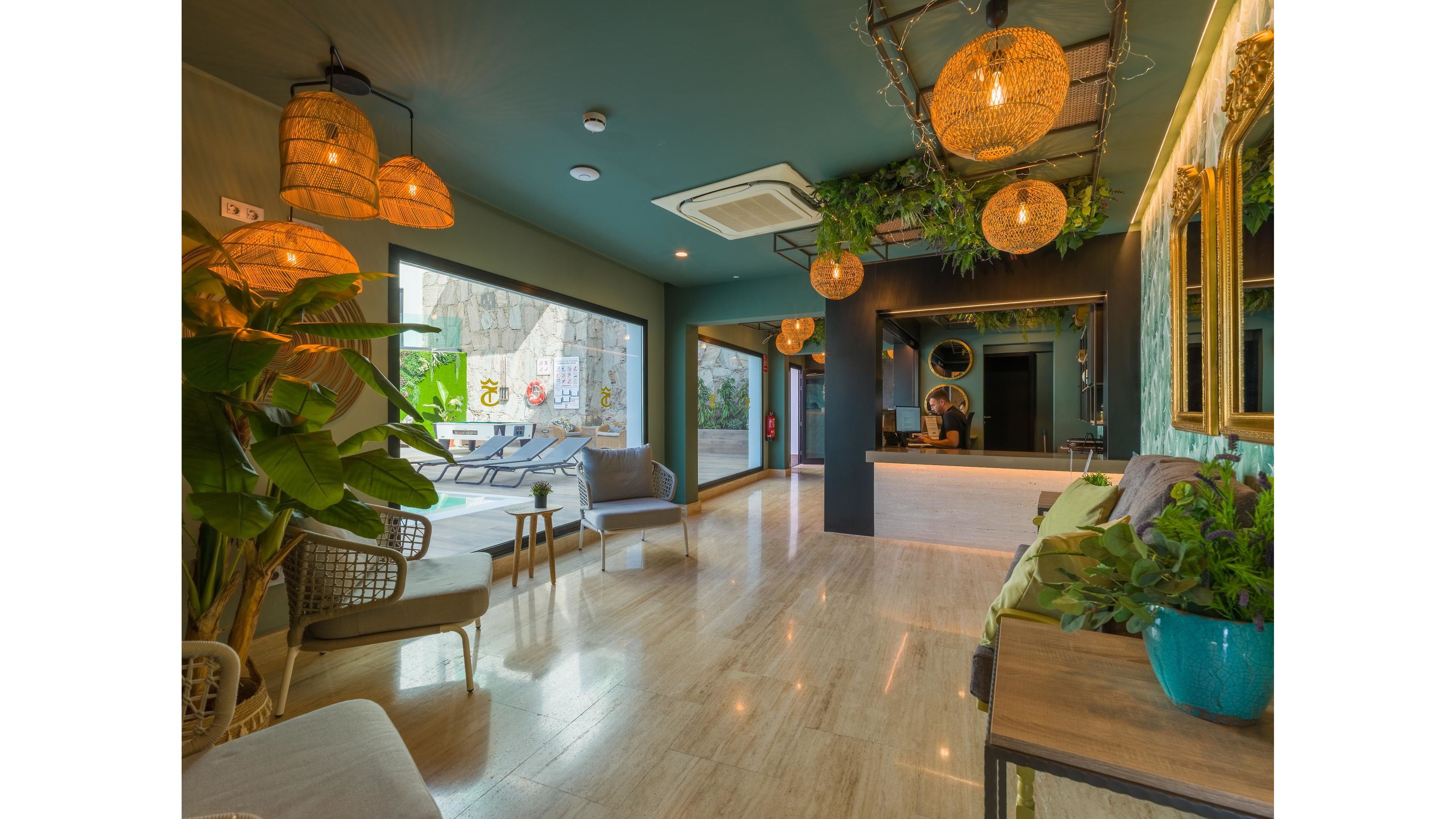 A contemporary hotel lobby features a reception desk and comfortable seating arranged along polished stone floors. Large open windows reveal an outdoor pool area with lounge chairs and greenery. Warm woven pendant lights and hanging plants create a tropical, inviting atmosphere. The space combines natural materials, teal walls, and decorative mirrors for a relaxed yet stylish interior.