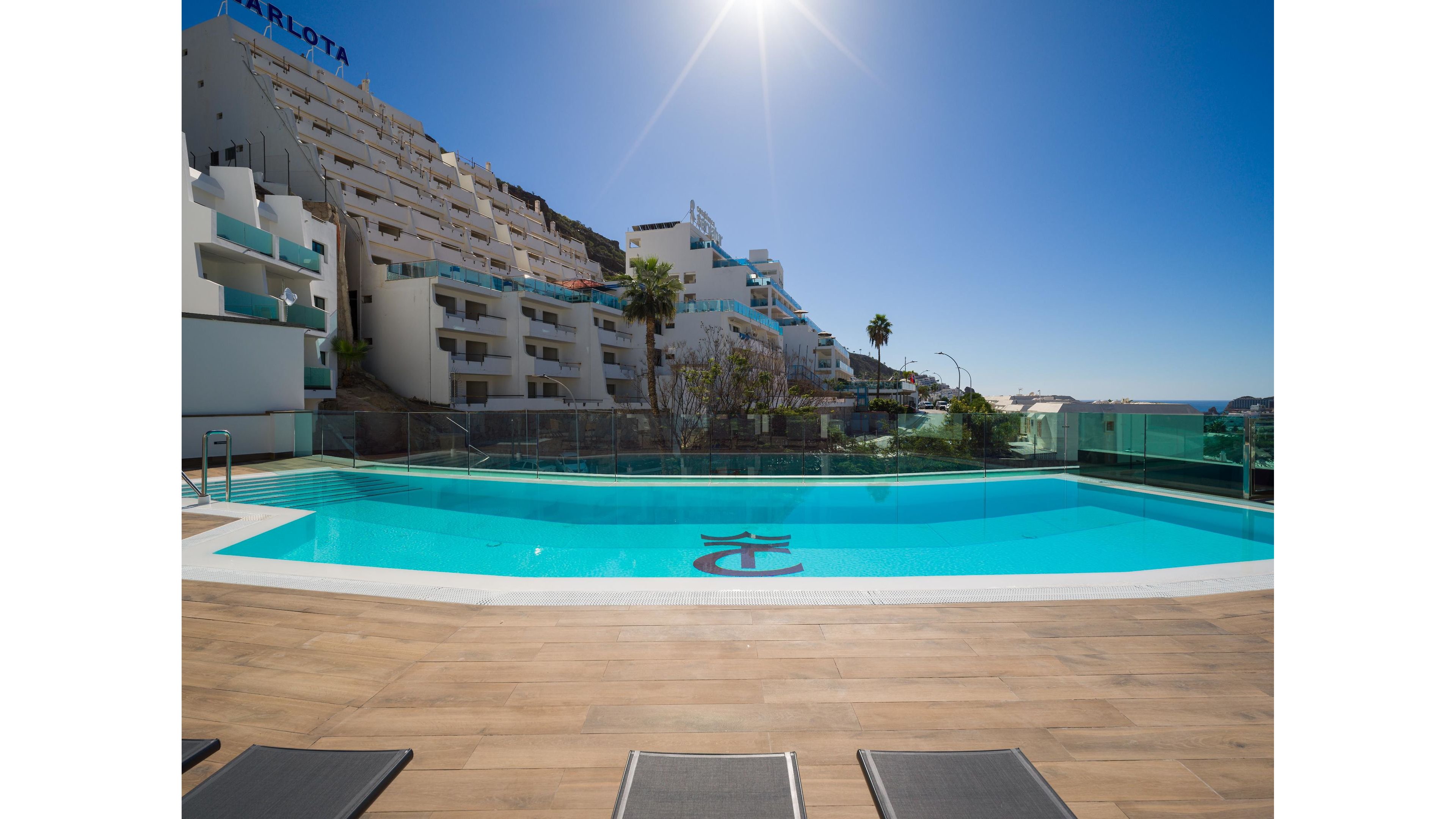 An outdoor swimming pool sits on a rooftop terrace overlooking a coastal town. Modern white hotel buildings rise on the hillside behind the pool under a clear blue sky. The sun shines brightly, casting reflections on the calm turquoise water and glass safety barriers. A few lounge chairs line the wooden deck, suggesting a relaxing vacation setting. Palm trees and distant sea views enhance the resort atmosphere.