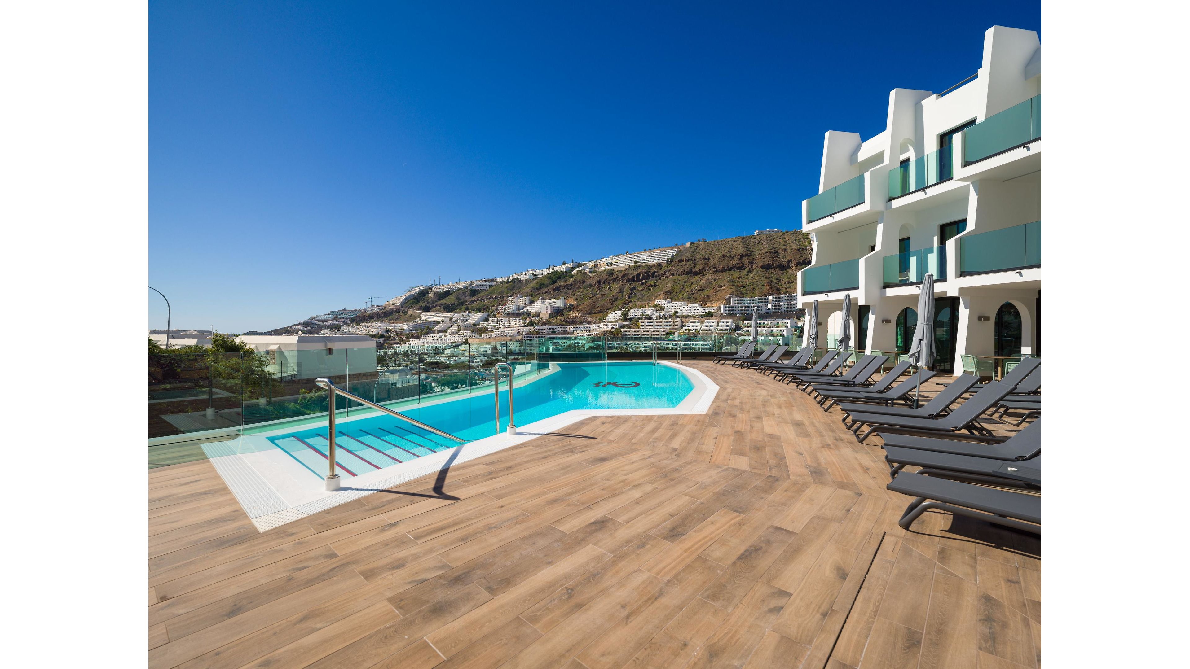 A modern hotel pool terrace overlooks a hillside town under a clear blue sky. The scene features a curved swimming pool surrounded by glass railings and a wide wooden deck. Numerous dark lounge chairs line the deck, all empty, suggesting a quiet and relaxing atmosphere. White contemporary buildings with green balcony panels frame the right side, while distant residences and rugged hills complete the coastal resort setting.