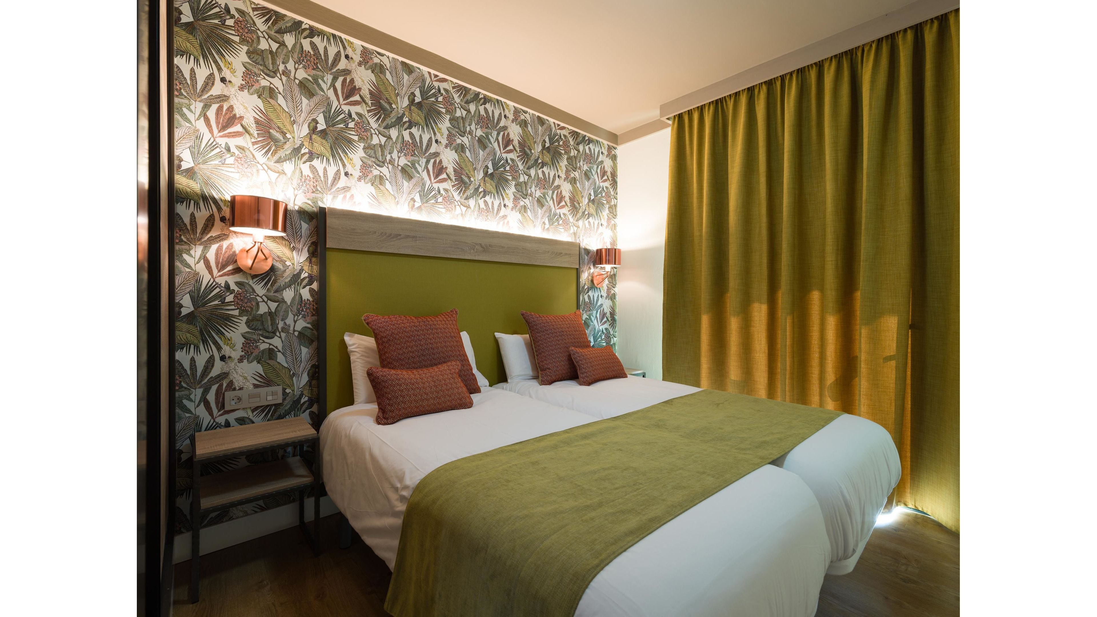 A contemporary hotel room features two twin beds pushed together with white linens and green bed runners. The headboard is upholstered in green fabric and framed by a wooden panel with soft backlighting. Patterned tropical wallpaper and matching green curtains add color and texture to the space. Warm wall-mounted lamps and decorative rust-colored pillows create a cozy, inviting atmosphere.