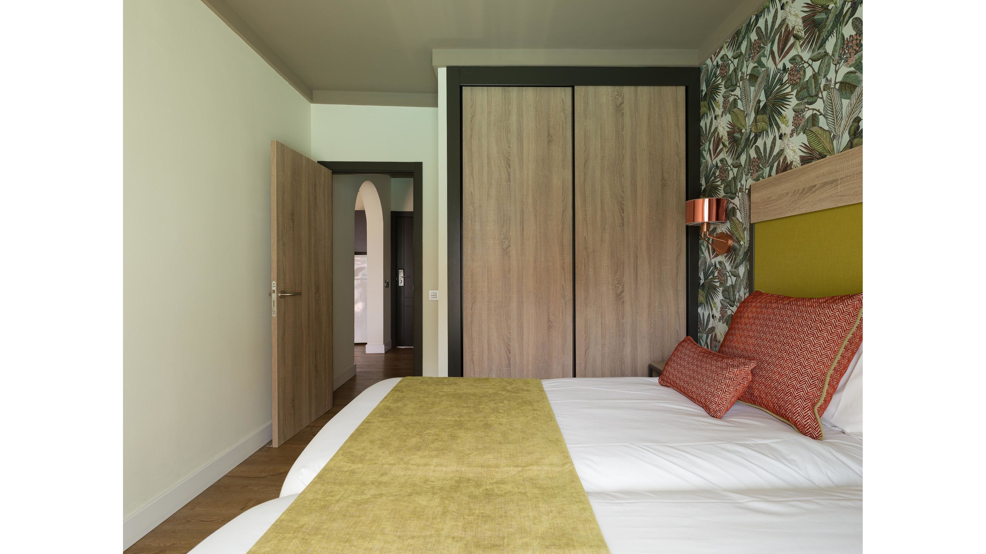 A contemporary bedroom features a neatly made bed with white linens, a green runner, and orange patterned pillows. Behind the bed, a tropical leaf-patterned accent wall adds visual interest and color. Opposite the bed, large wooden sliding closet doors provide storage and a minimalist look. An open door reveals a hallway with an arched doorway, contributing to the room’s airy and inviting atmosphere.