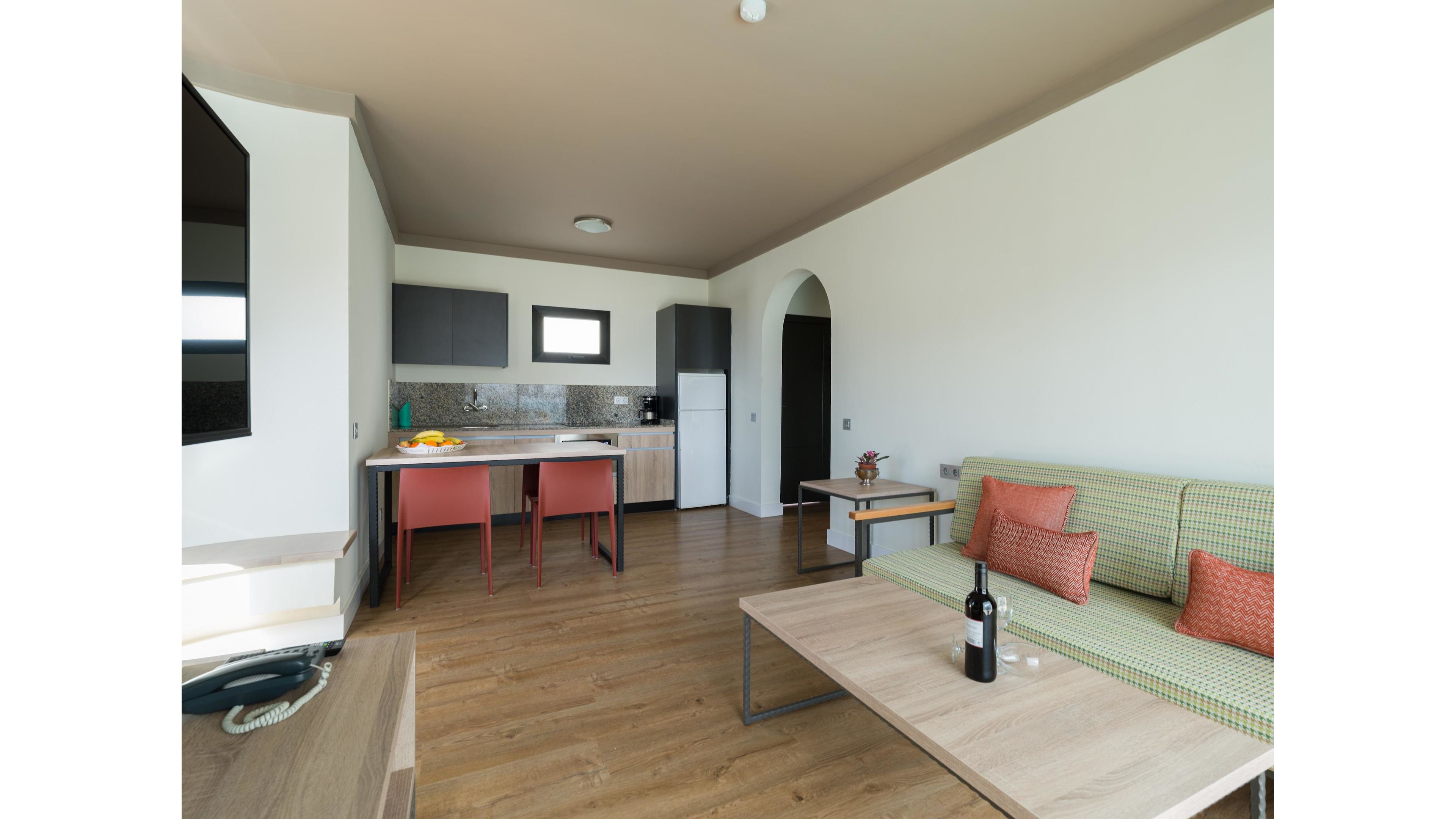 A contemporary open-plan living space combines a small kitchen and a cozy seating area. The kitchen features light wood cabinets, a tiled backsplash, and a compact dining table with two red chairs. In the foreground, a green sofa with orange cushions faces a wooden coffee table holding a wine bottle and glasses. Neutral walls and a wooden floor create a clean, minimalist atmosphere with soft natural light.