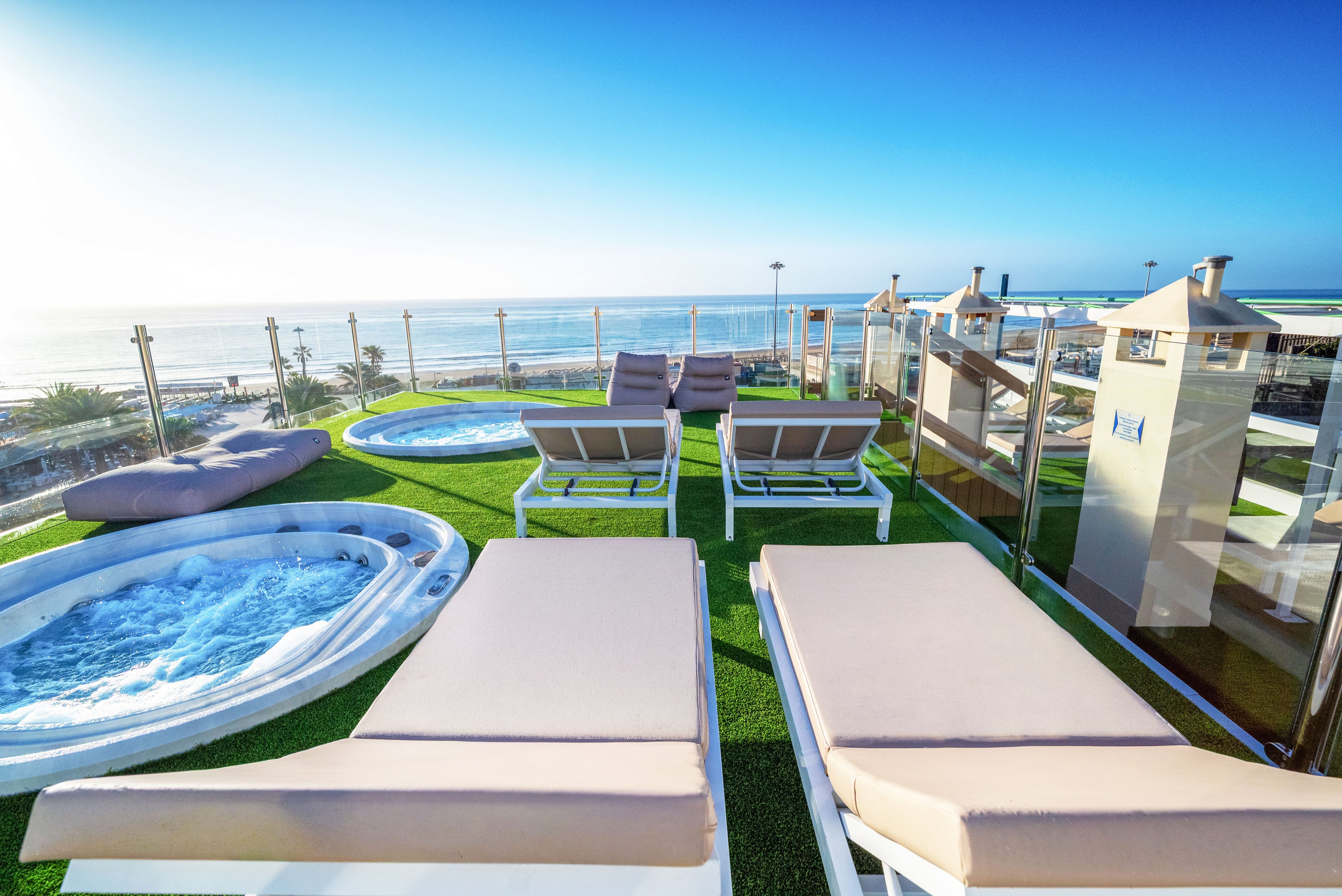 A modern rooftop terrace features cushioned sun loungers facing the ocean. Two circular jacuzzis are set into artificial grass, with clear glass railings framing the coastal view. The bright blue sky and calm sea create a relaxing vacation atmosphere. The scene suggests a resort or spa environment designed for leisure and sunbathing.