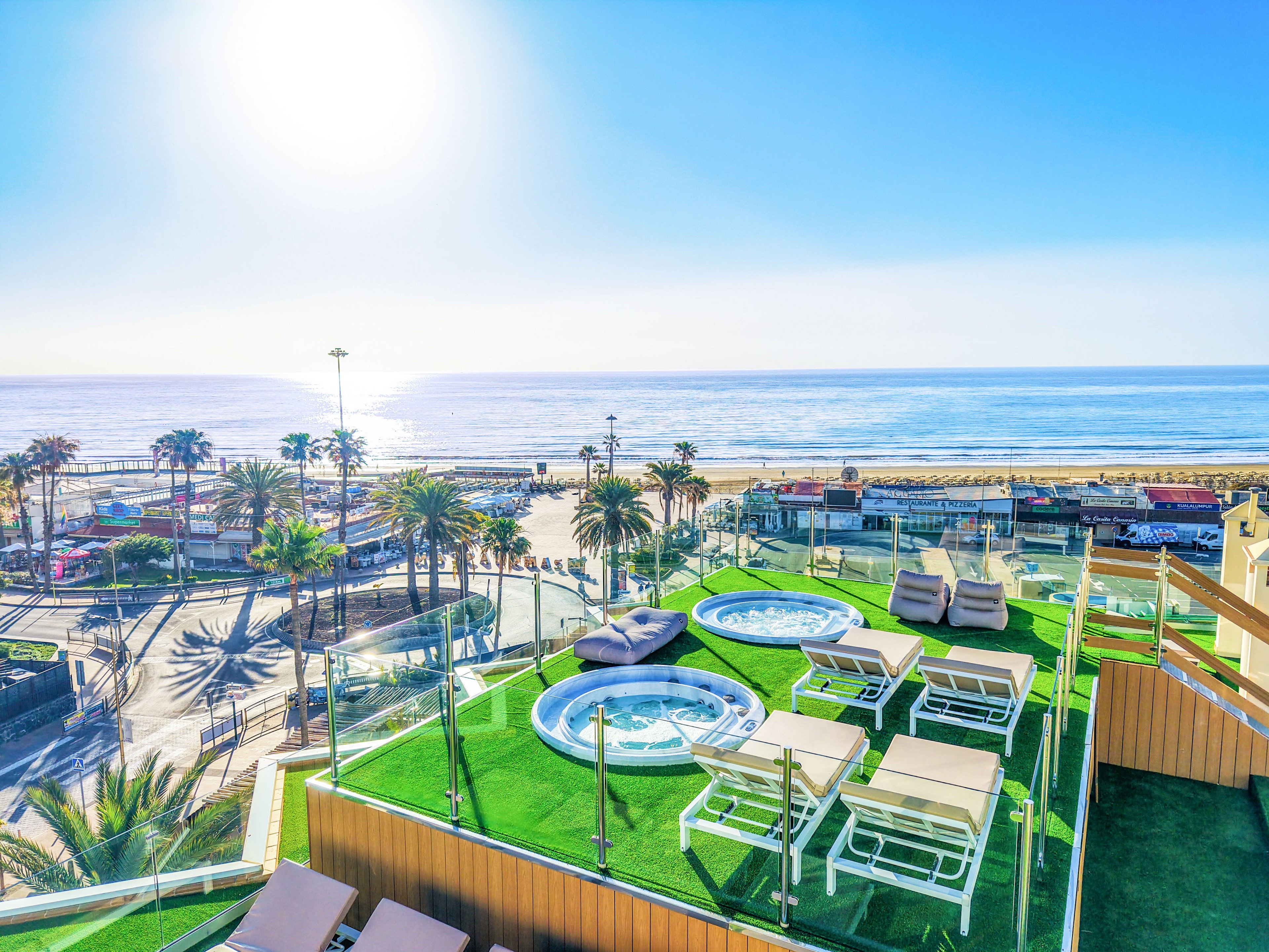 A modern rooftop terrace features multiple jacuzzis and sun loungers on artificial grass, enclosed by glass railings. Below, a lively seaside promenade with palm trees and parked cars stretches along the coastline. The bright sun reflects off the calm ocean, creating a vibrant, summery atmosphere. The scene suggests a relaxing vacation spot with panoramic views of the beach and waterfront.