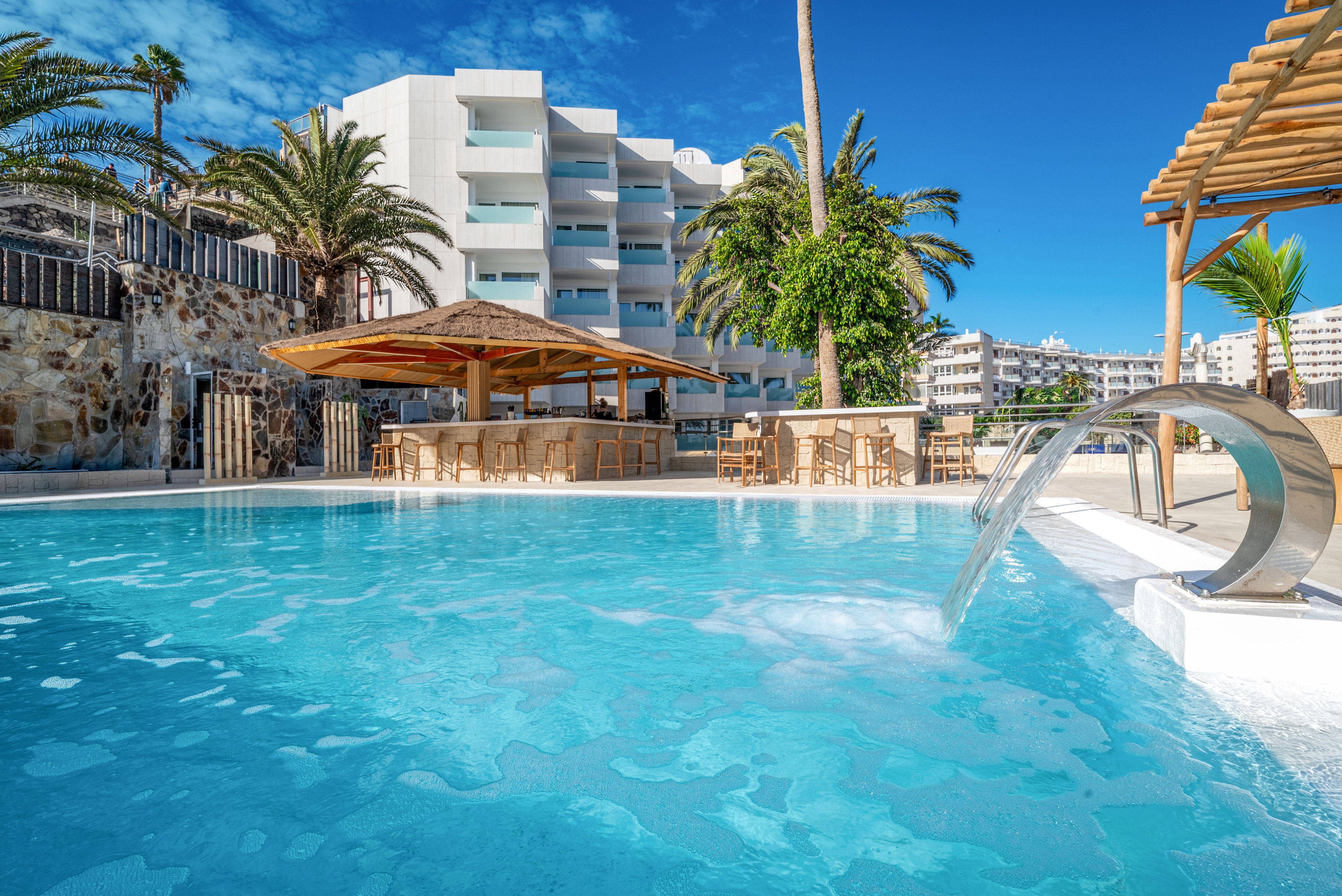 A clear blue resort swimming pool is shown in the foreground with a modern metal water spout cascading into the water. Behind the pool, there is a shaded bar area with wooden stools and a pergola structure. White multi-story hotel buildings and lush palm trees frame the scene under a bright blue sky. The image conveys a sunny, relaxing vacation atmosphere at a coastal or tropical destination.