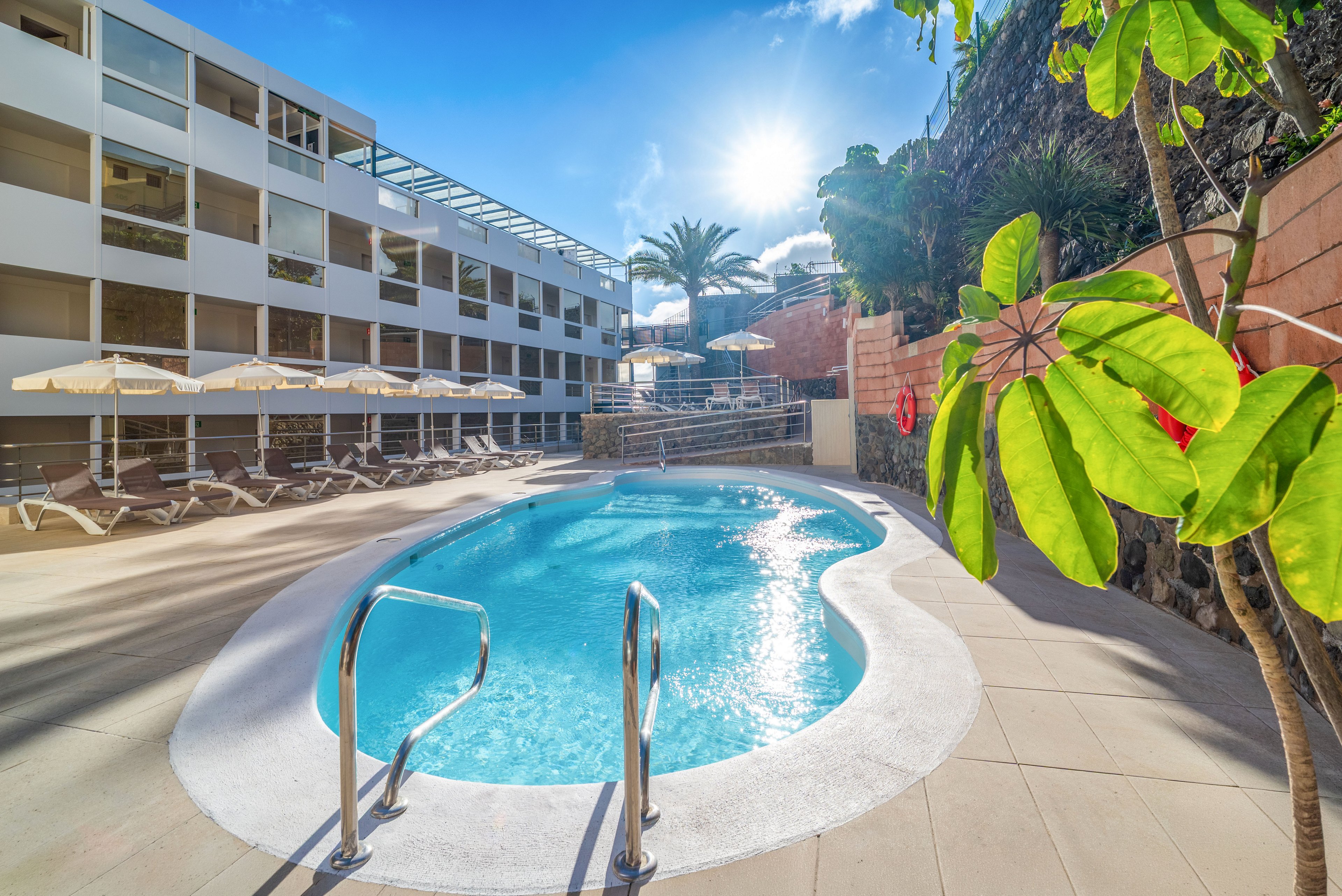A bright outdoor swimming pool sits in the courtyard of a modern hotel or apartment complex. Sun loungers and parasols line the poolside terrace, suggesting a relaxing vacation setting. The sun shines strongly from a clear blue sky, casting reflections on the water and surrounding buildings. Tropical plants and a rocky wall add a touch of greenery and natural texture to the scene.