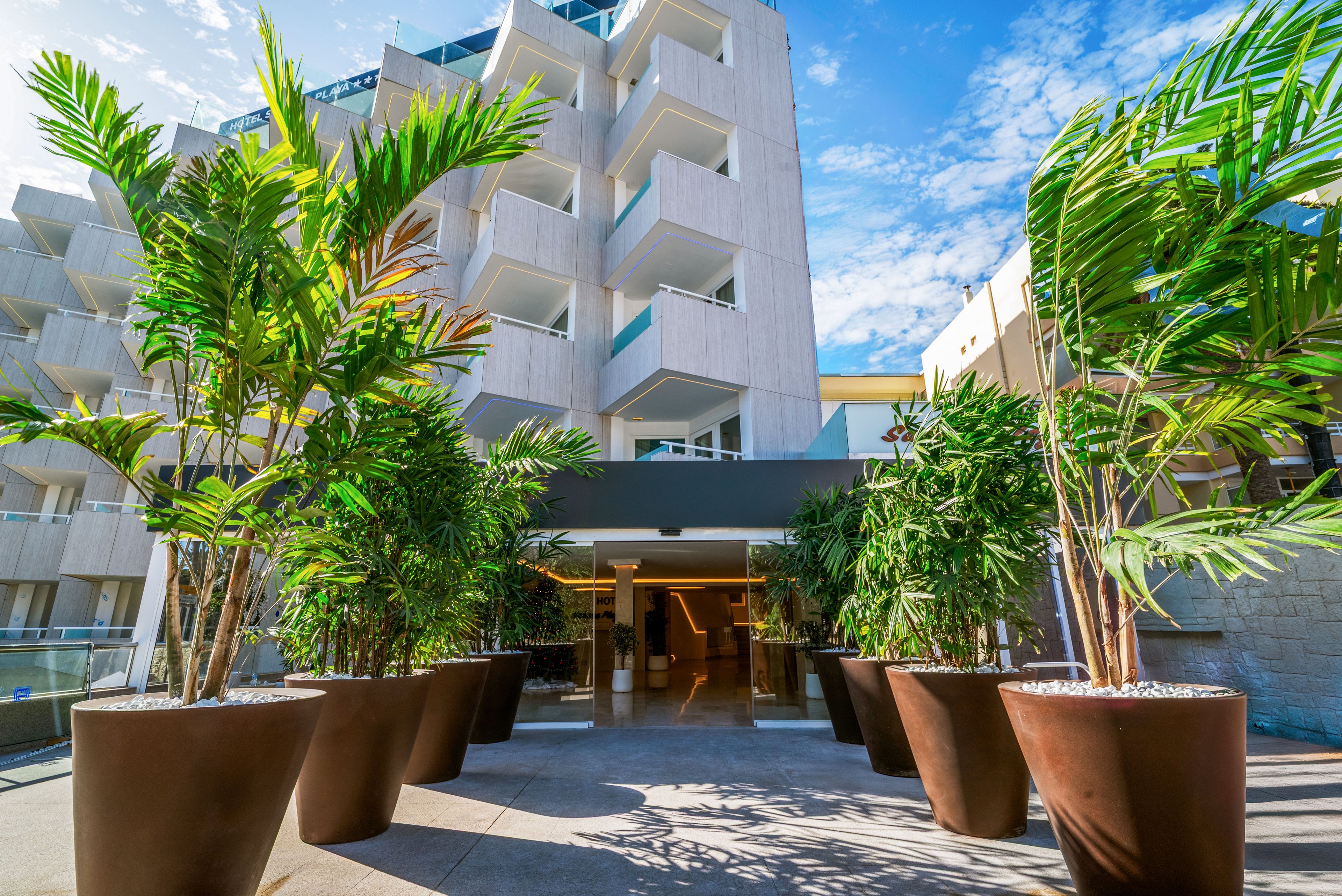 A contemporary multi-story hotel building features an inviting entrance lined with large potted tropical plants. The architecture showcases clean lines, balconies, and light-colored facades under a bright blue sky with scattered clouds. The wide walkway leads guests toward the lobby area, framed by greenery on both sides. The overall scene conveys a modern, resort-like atmosphere in a sunny outdoor setting.