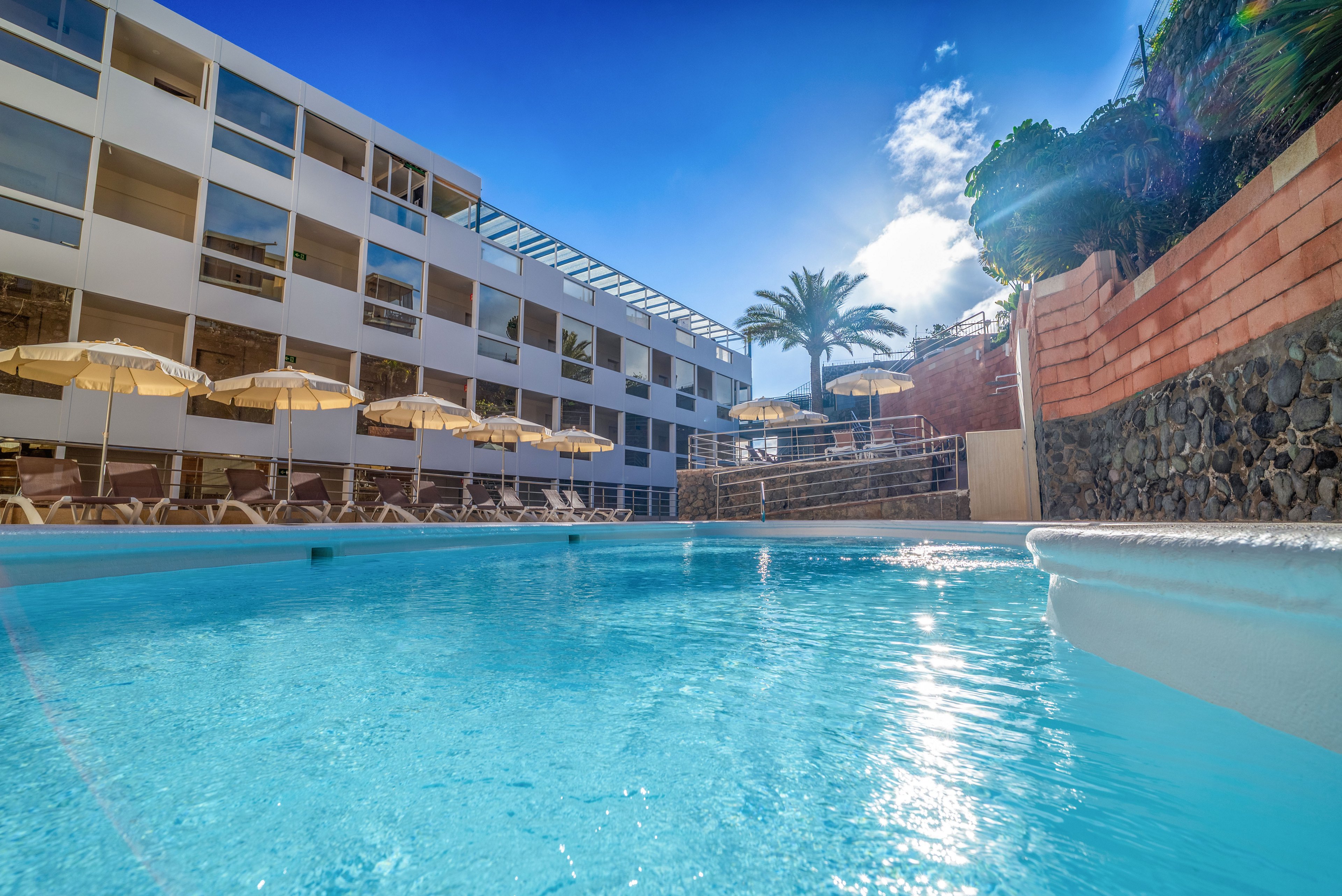 A clear blue outdoor swimming pool sits in front of a modern multi-story hotel building. Sun loungers and cream-colored umbrellas line the poolside terrace, suggesting a relaxing vacation setting. Palm trees and stone walls frame the area, while bright sunlight and a blue sky create a warm, inviting atmosphere. The scene conveys leisure, resort comfort, and sunny holiday vibes.